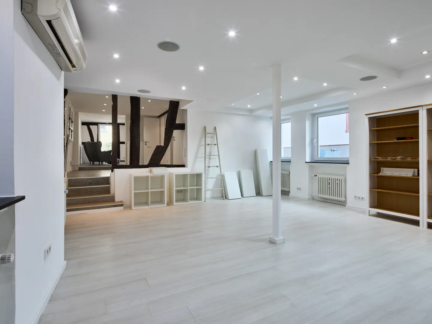 Bright, empty room with white walls, light wood floors, and recessed lighting. A staircase leads to a raised area with dark wood beams.
