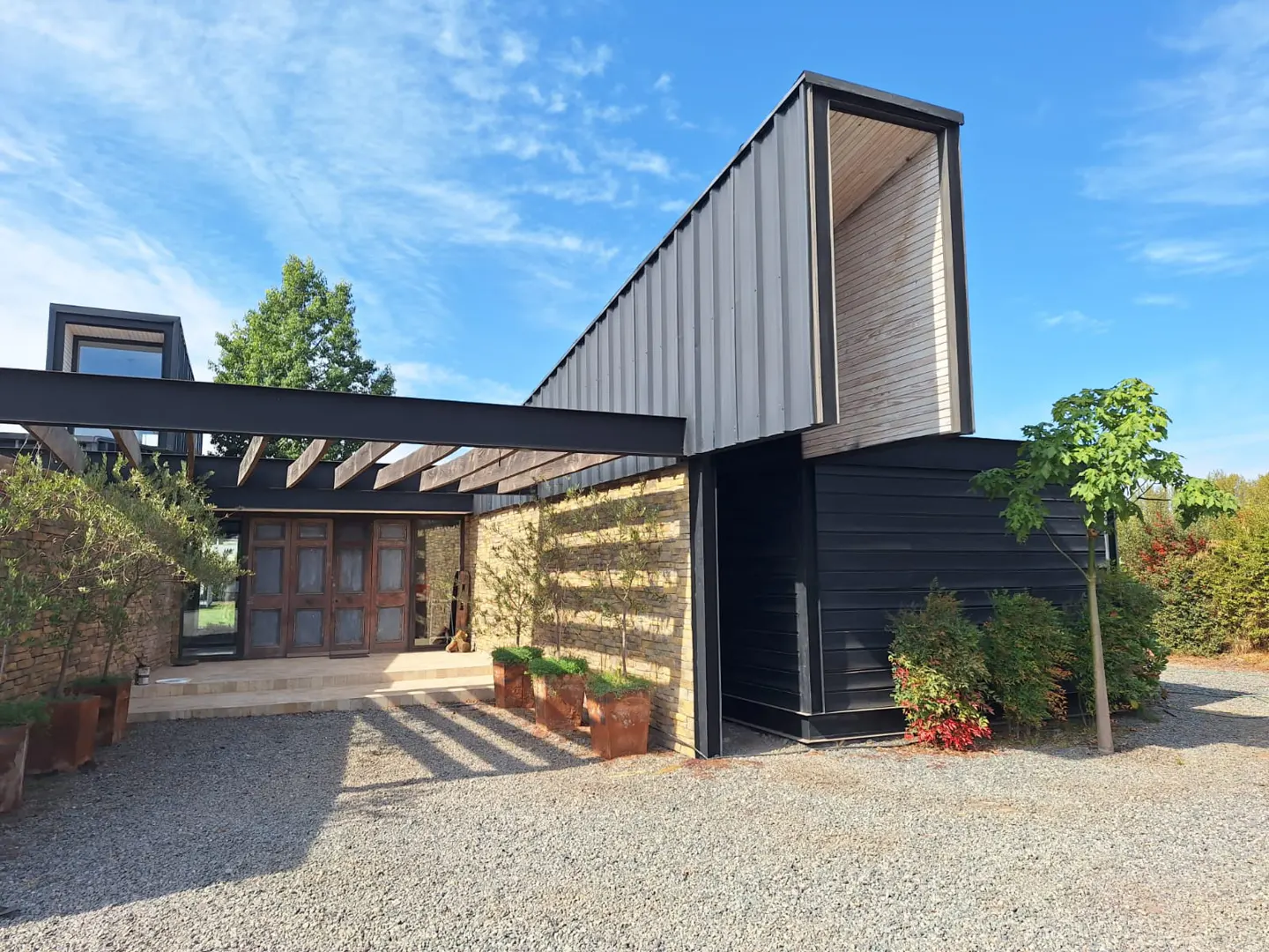 Modern home exterior with a black metal and wood design, stone accents, and a gravel driveway under a blue sky.