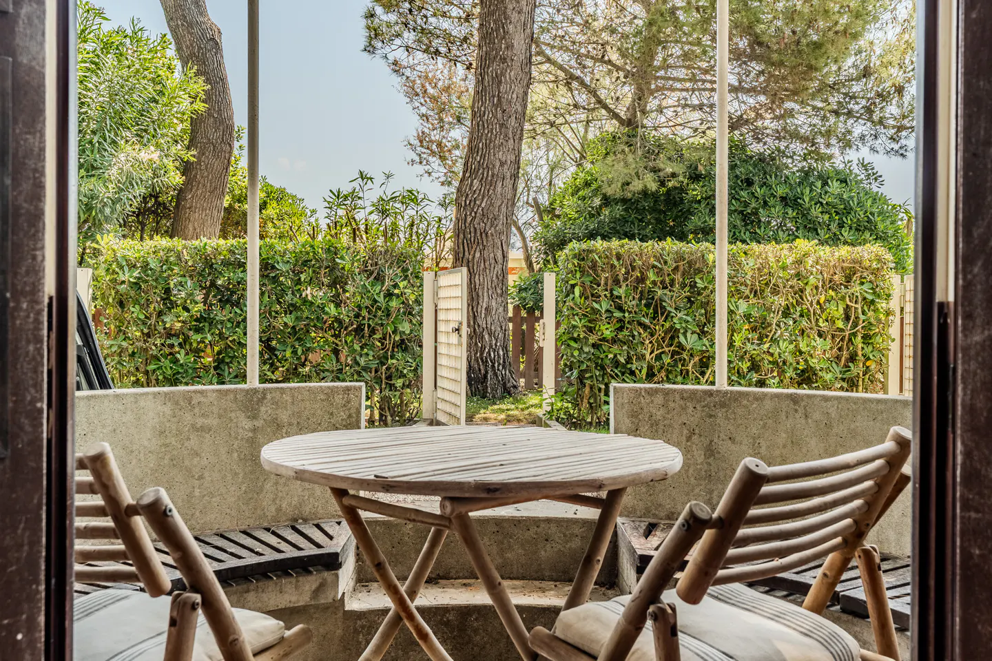 View from doorway of patio with round wooden table and chairs. Green hedges and trees in background.