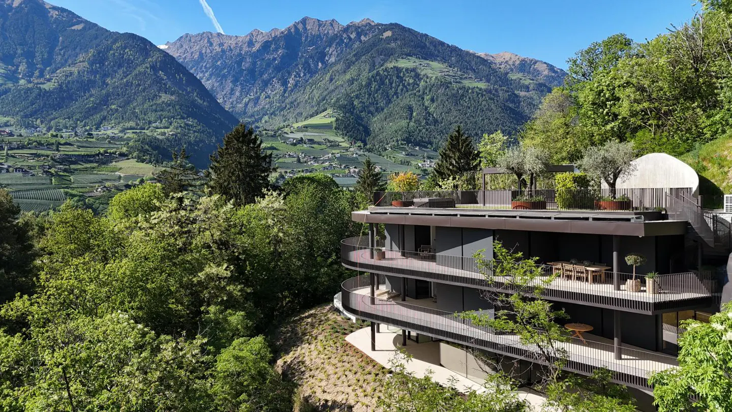 Modern three-story home with curved balconies, nestled in lush green trees with mountains in the background.