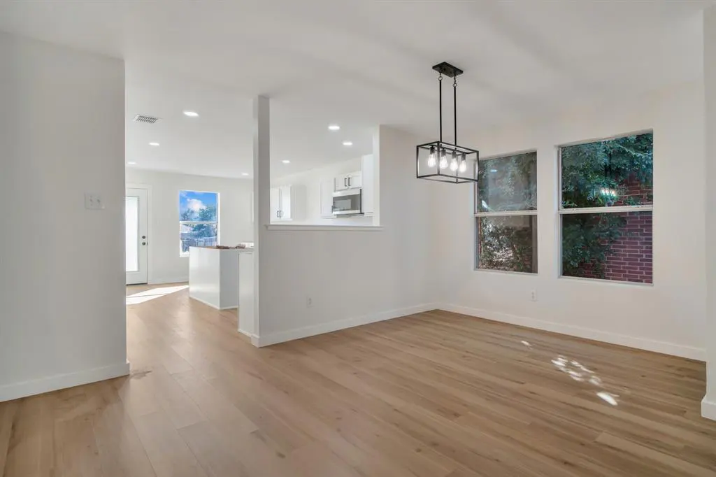 Bright, open-concept dining area with wood floors, white walls, and a modern black chandelier. Windows offer views of lush greenery.
