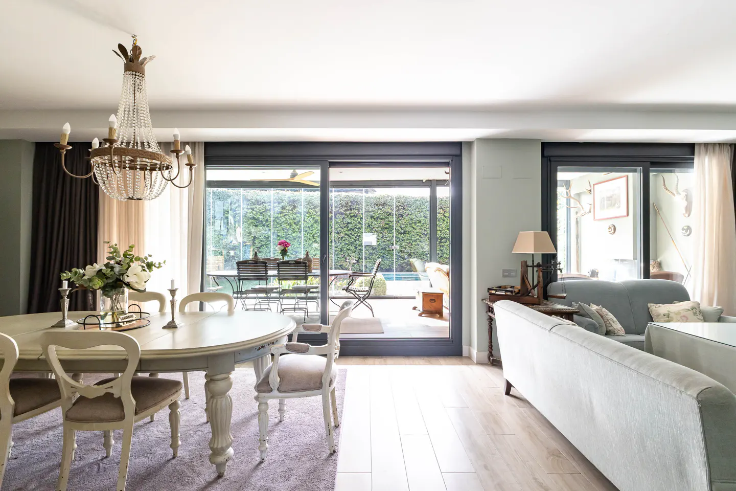 Bright living room with a white dining table, chandelier, and a view of a green garden through sliding glass doors.