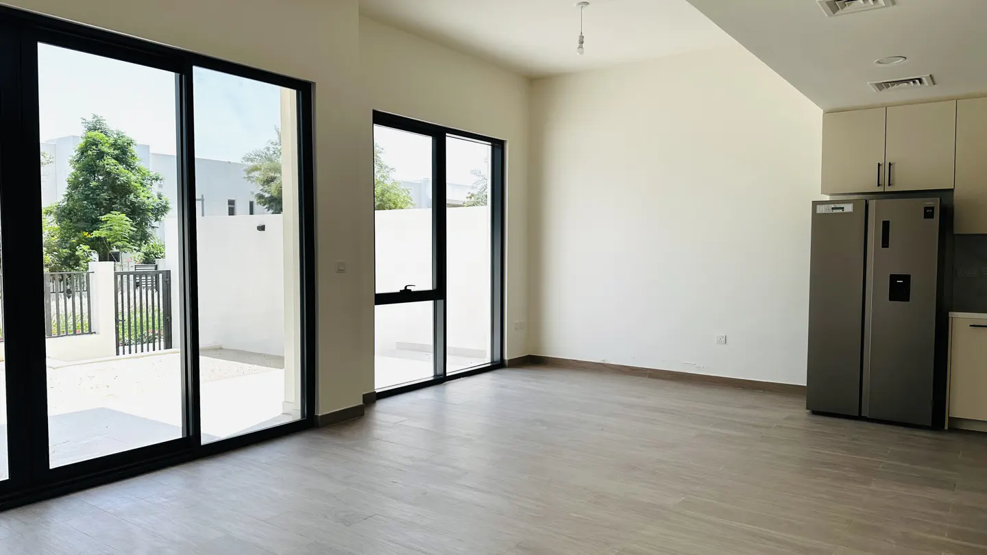 An empty room with light wood floors, white walls, and black-framed sliding glass doors to the outside. A stainless steel refrigerator is on the right.