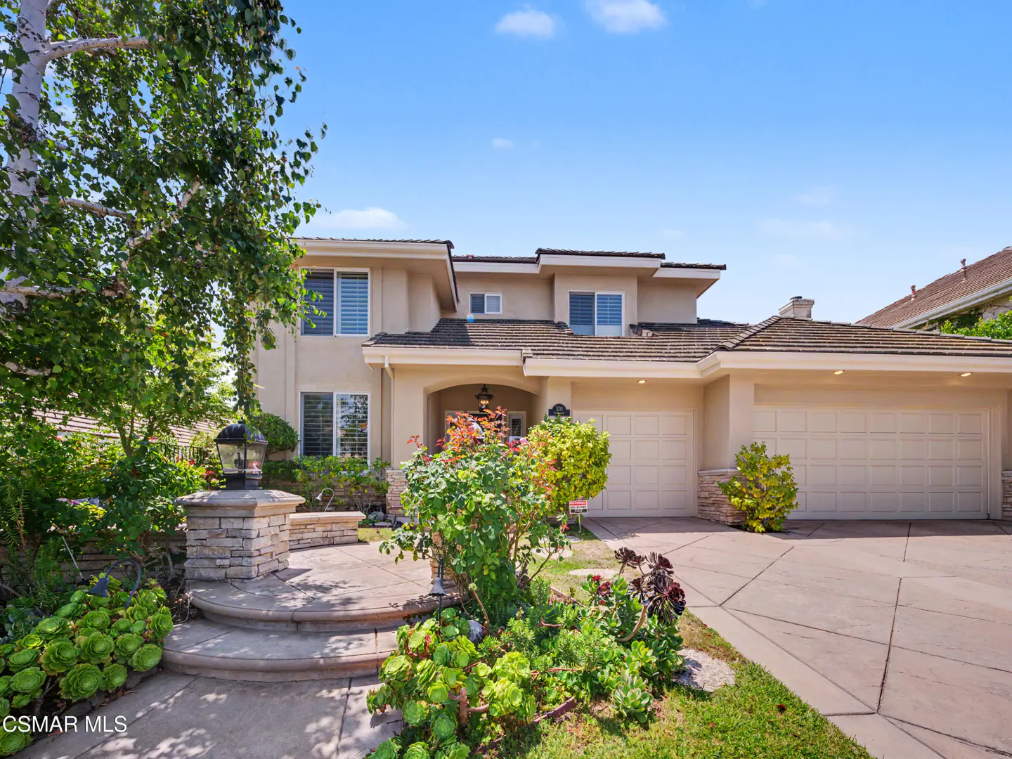 Two-story beige suburban home with a multi-car garage, stone-accented entryway, and lush front yard landscaping.