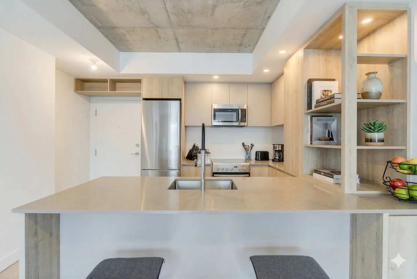 Modern kitchen with stainless steel appliances, light wood cabinets, and a concrete ceiling. A large island with a sink and two gray stools is in the foreground.
