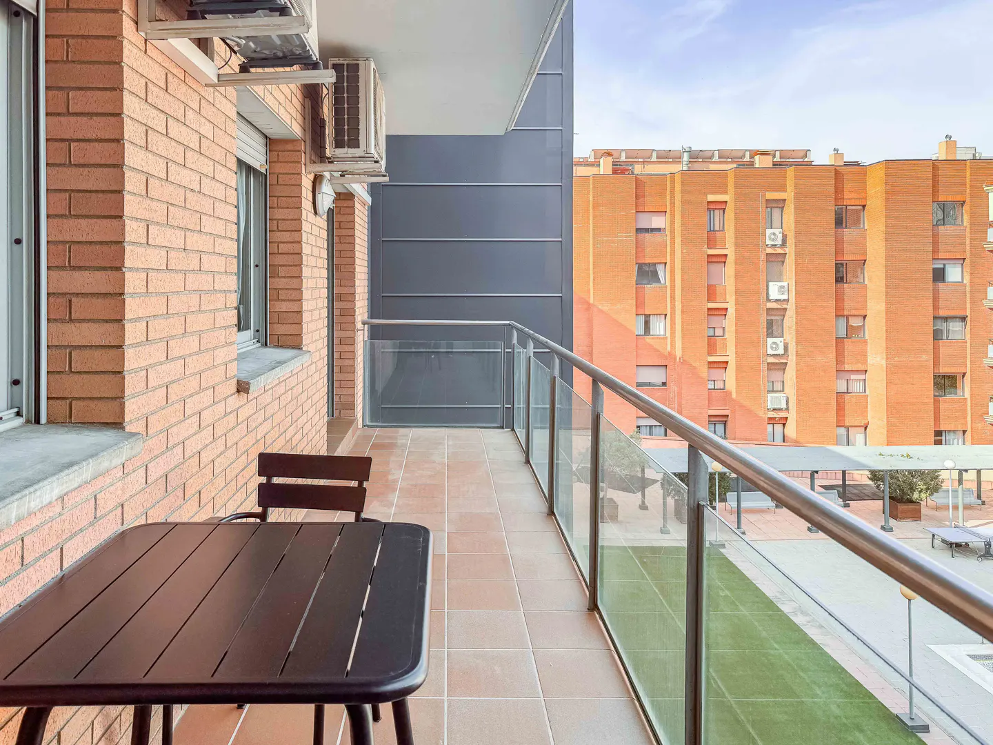 Balcony with a dark table and chair, brick wall, and glass railing overlooking a courtyard with a brick building in the background.