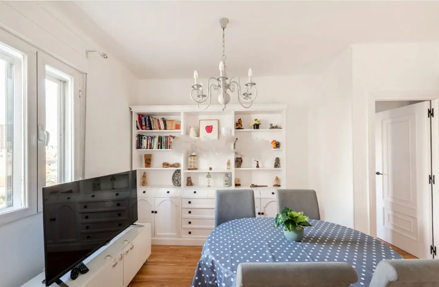 Bright dining room with white walls, wood floors, and a large white bookcase. A table with a blue polka dot tablecloth and gray chairs sits in the center. A TV is on the left.