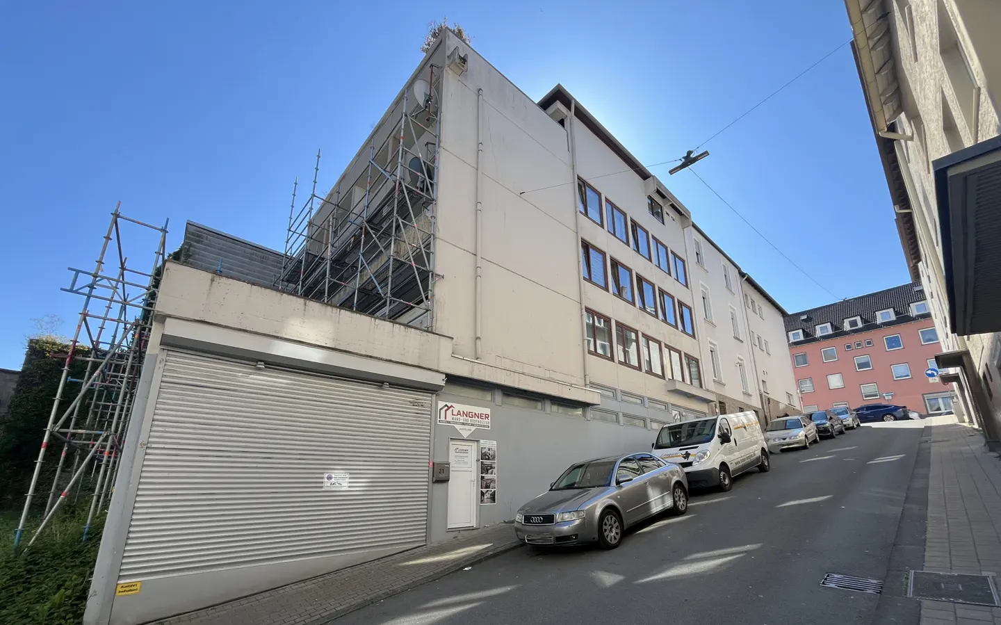 Exterior view of a building with scaffolding, cars parked on a street, and a blue sky.