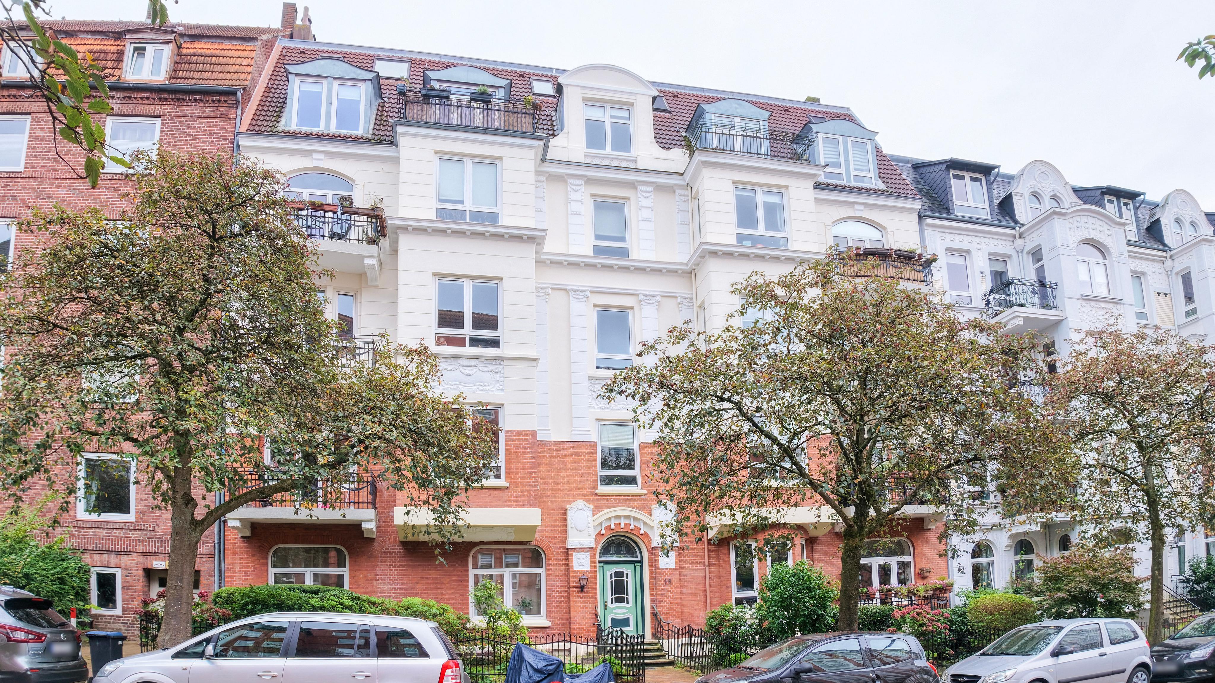 A multi-story European apartment building with a red brick base, white upper facade, and several balconies on a tree-lined street. [AI-generated text]