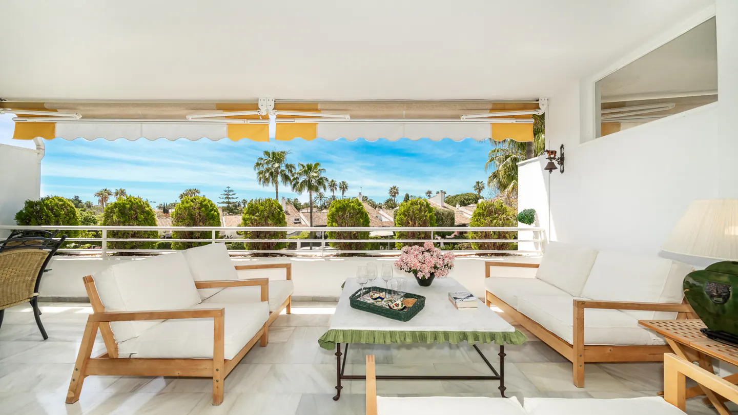 Bright balcony with white sofas, a marble table with flowers, and a view of palm trees under a blue sky.