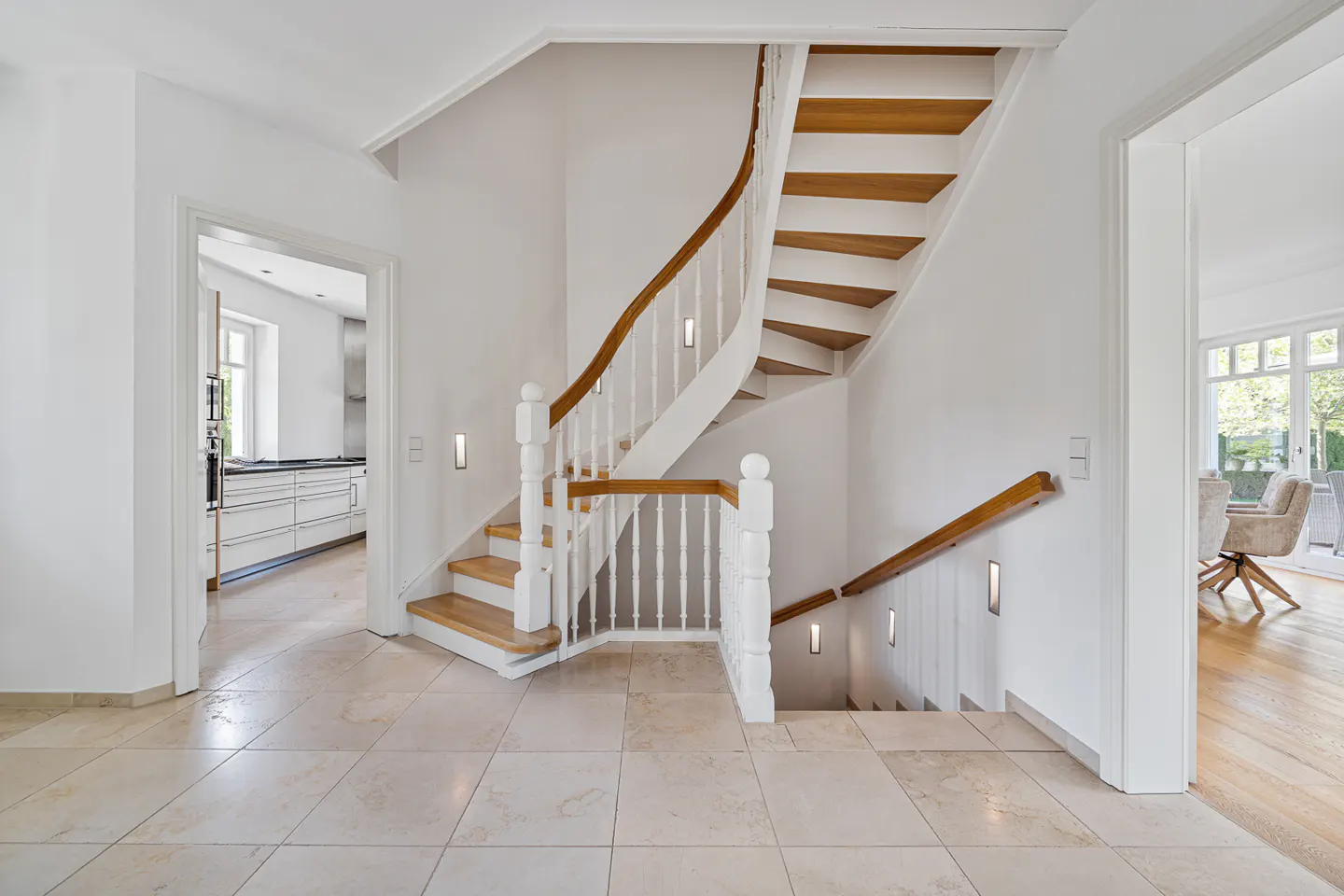 Bright foyer with a winding staircase. White walls, wood steps, and tile floor. Kitchen and dining room visible through doorways.