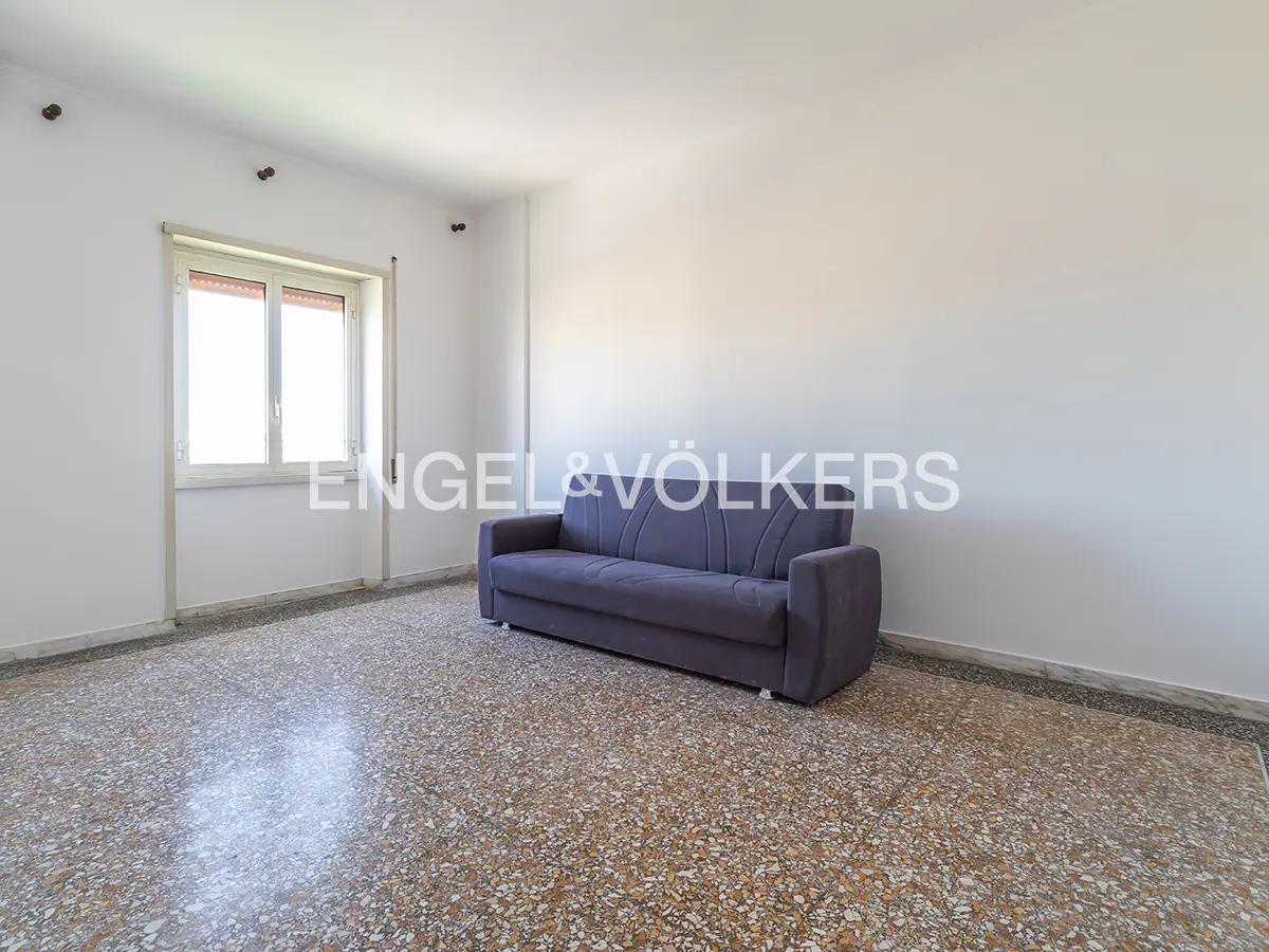 A bright, empty room with a blue sofa, terrazzo floor, and a window letting in natural light.