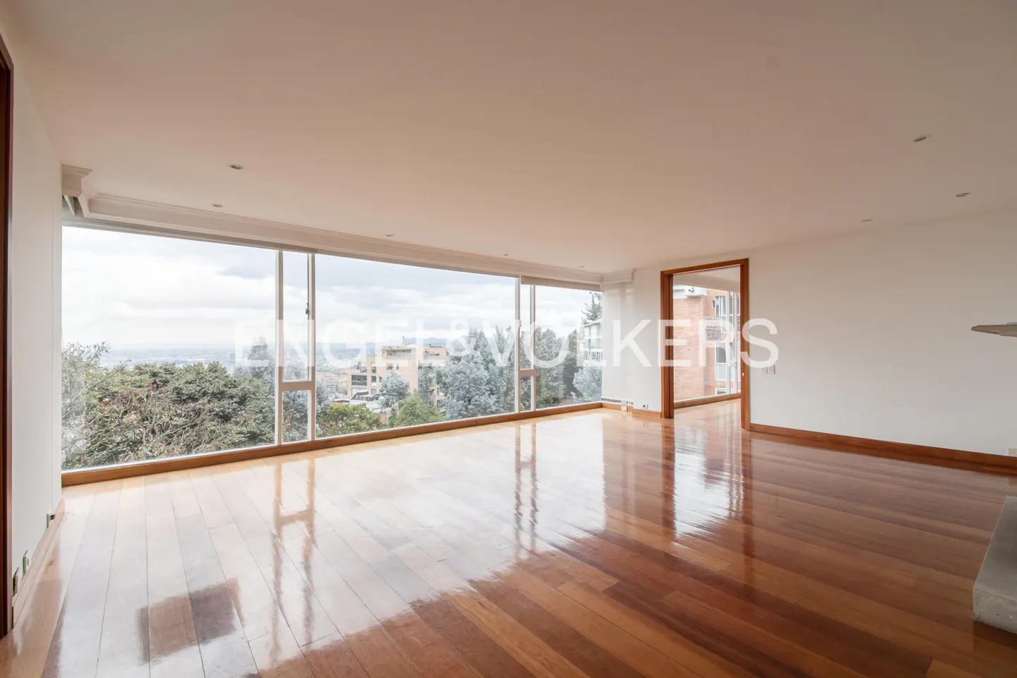Bright, empty living room with shiny wood floors and a large window showing a city view. White walls and a doorway are visible.