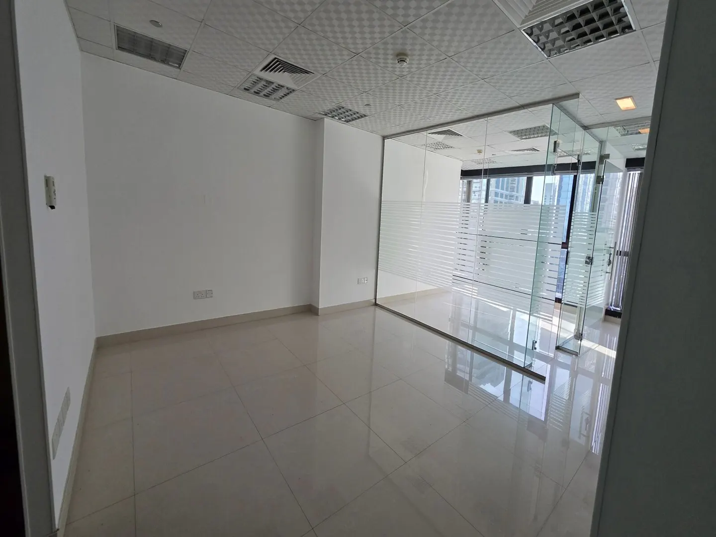Empty office space with white walls, tiled floor, and glass-walled room with frosted stripes.