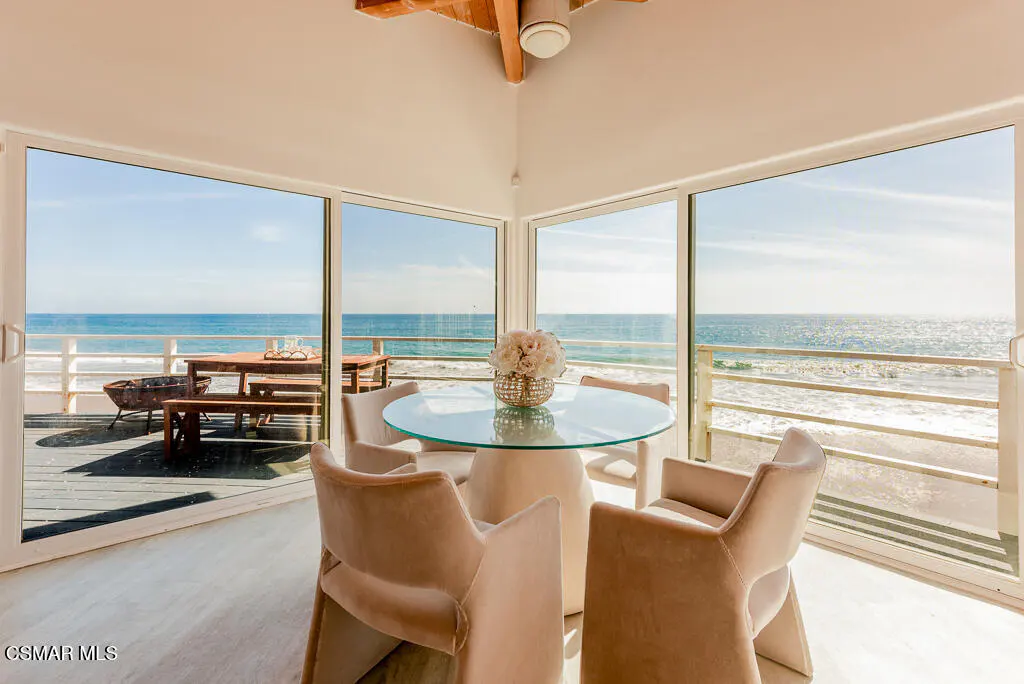 Bright dining room with ocean view. Glass table with beige chairs. Deck with table, benches, and fire pit visible through sliding glass doors.