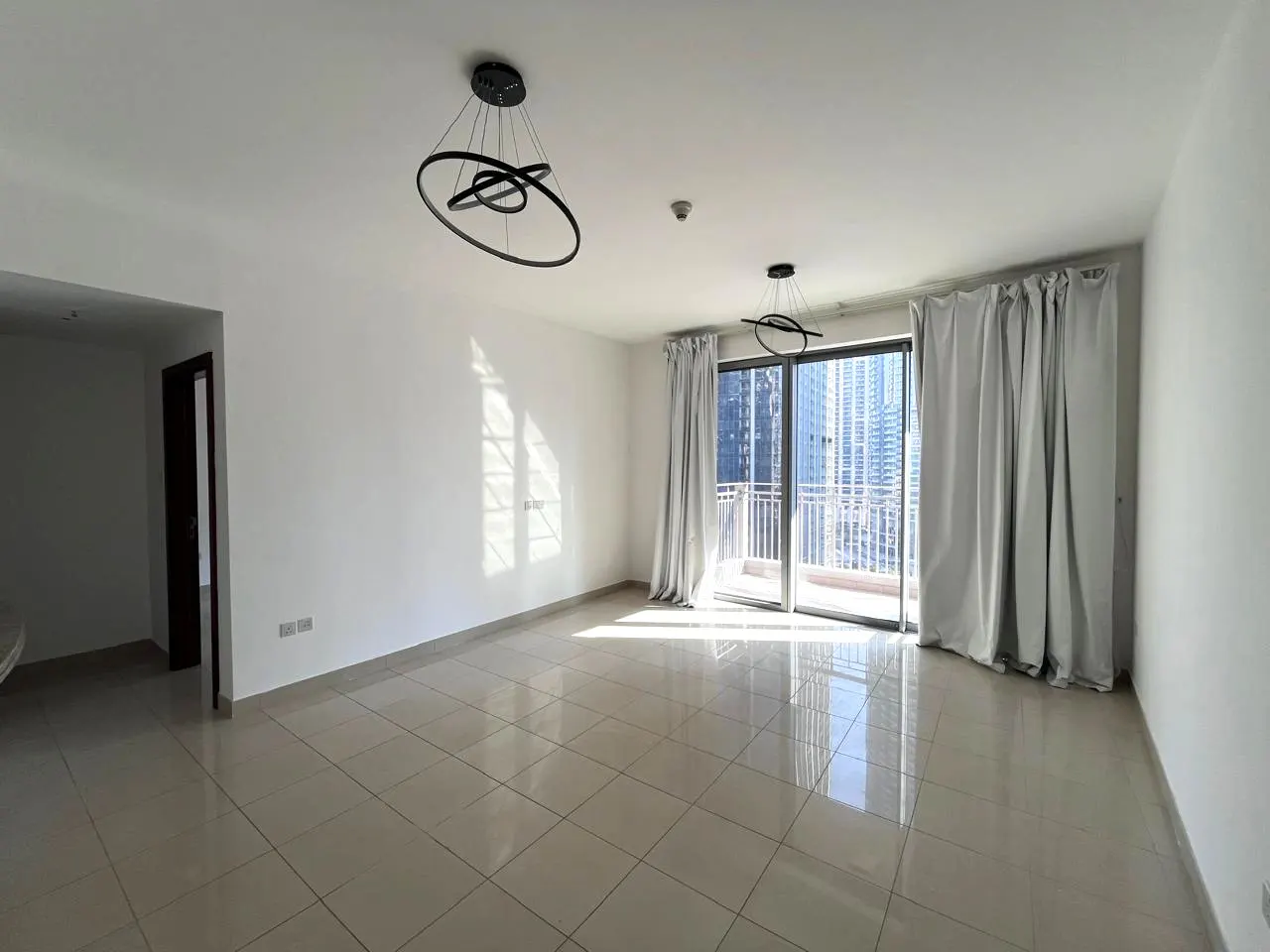 Bright, empty room with white walls, tile floor, and balcony with city view. Modern black light fixtures hang from the ceiling.
