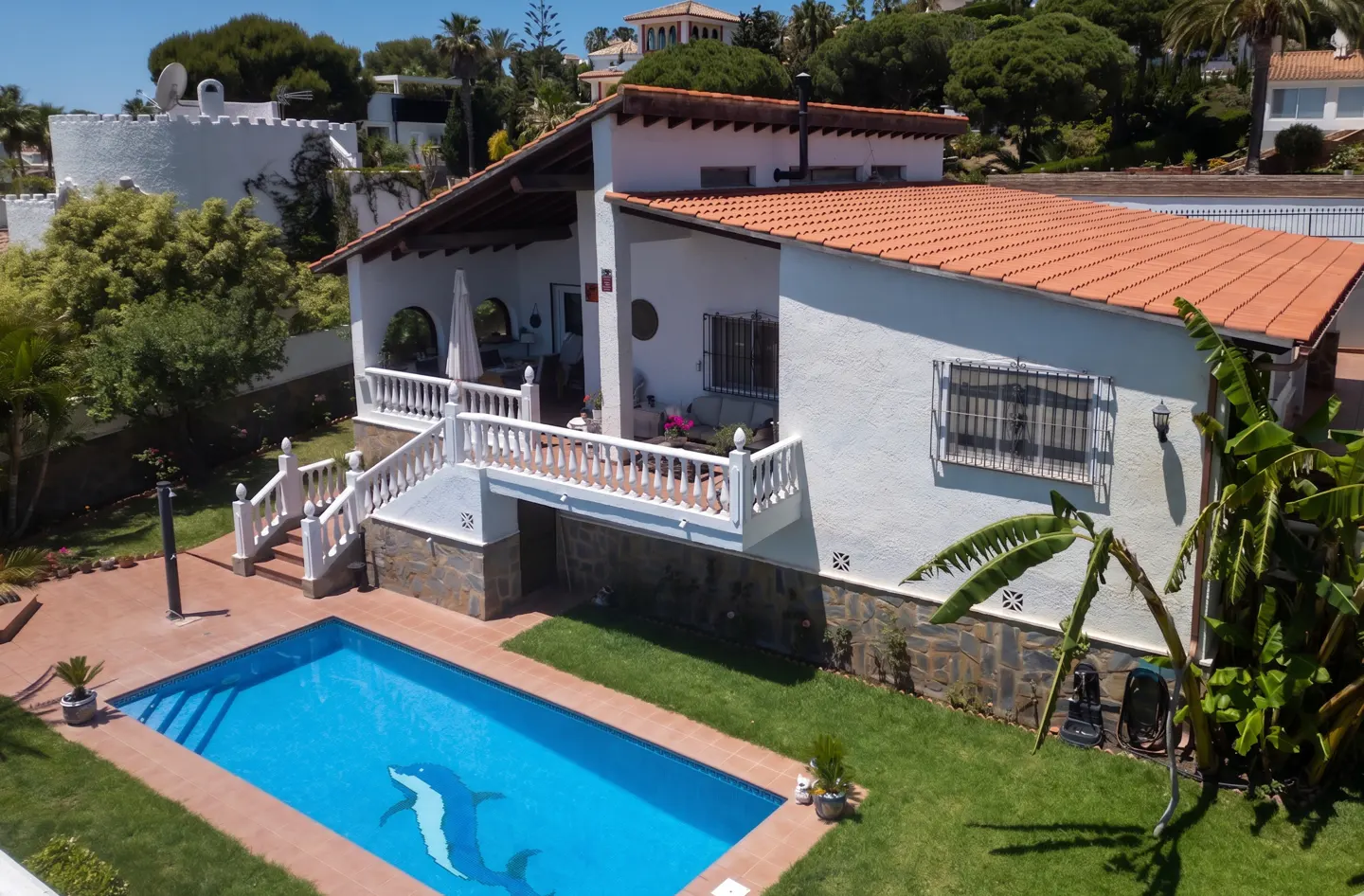 Aerial view of a white house with a red tile roof, a blue pool with a dolphin mosaic, and green grass.
