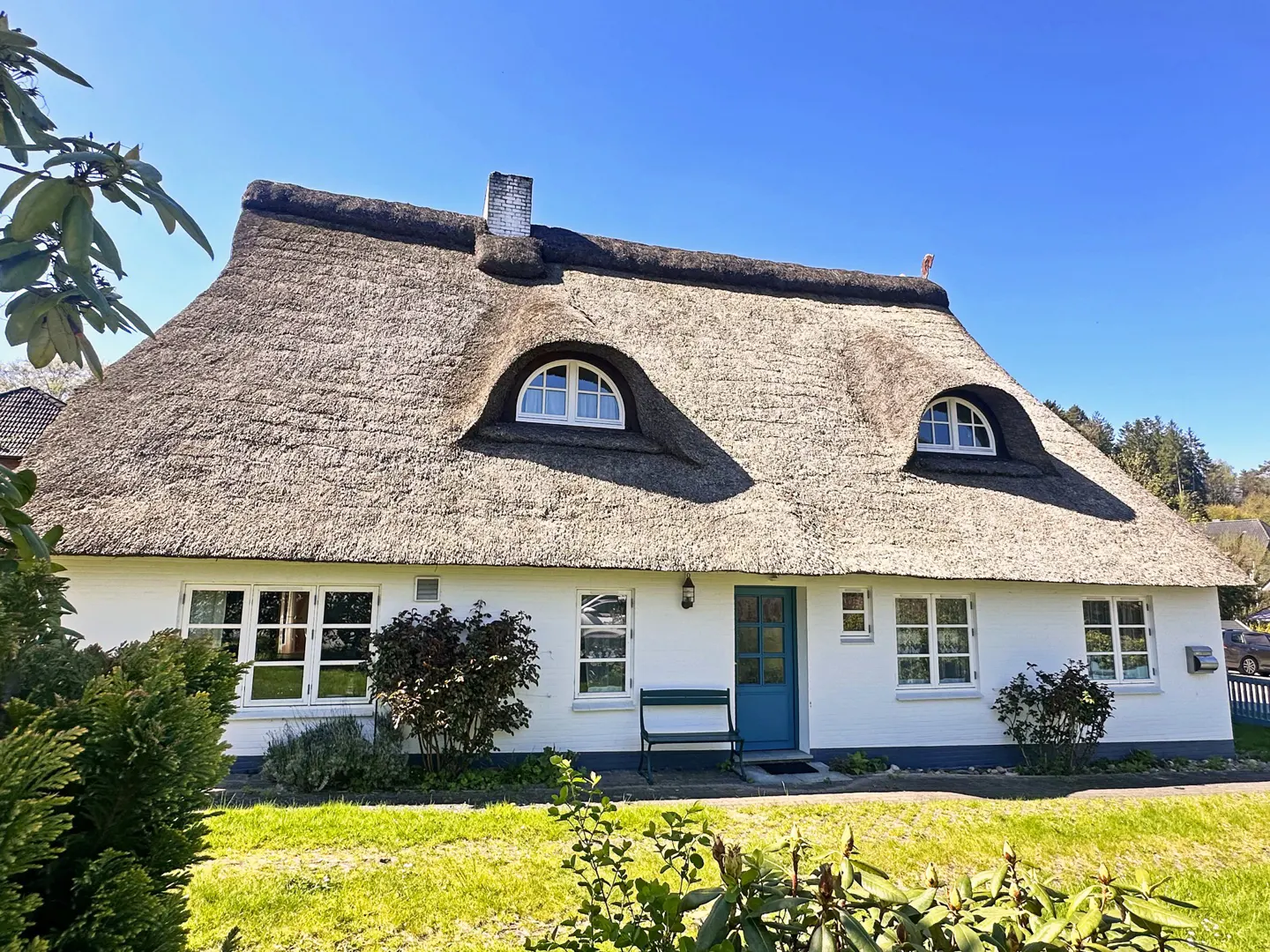 Charming white cottage with a thatched roof, dormer windows, and a blue front door, surrounded by a green lawn.