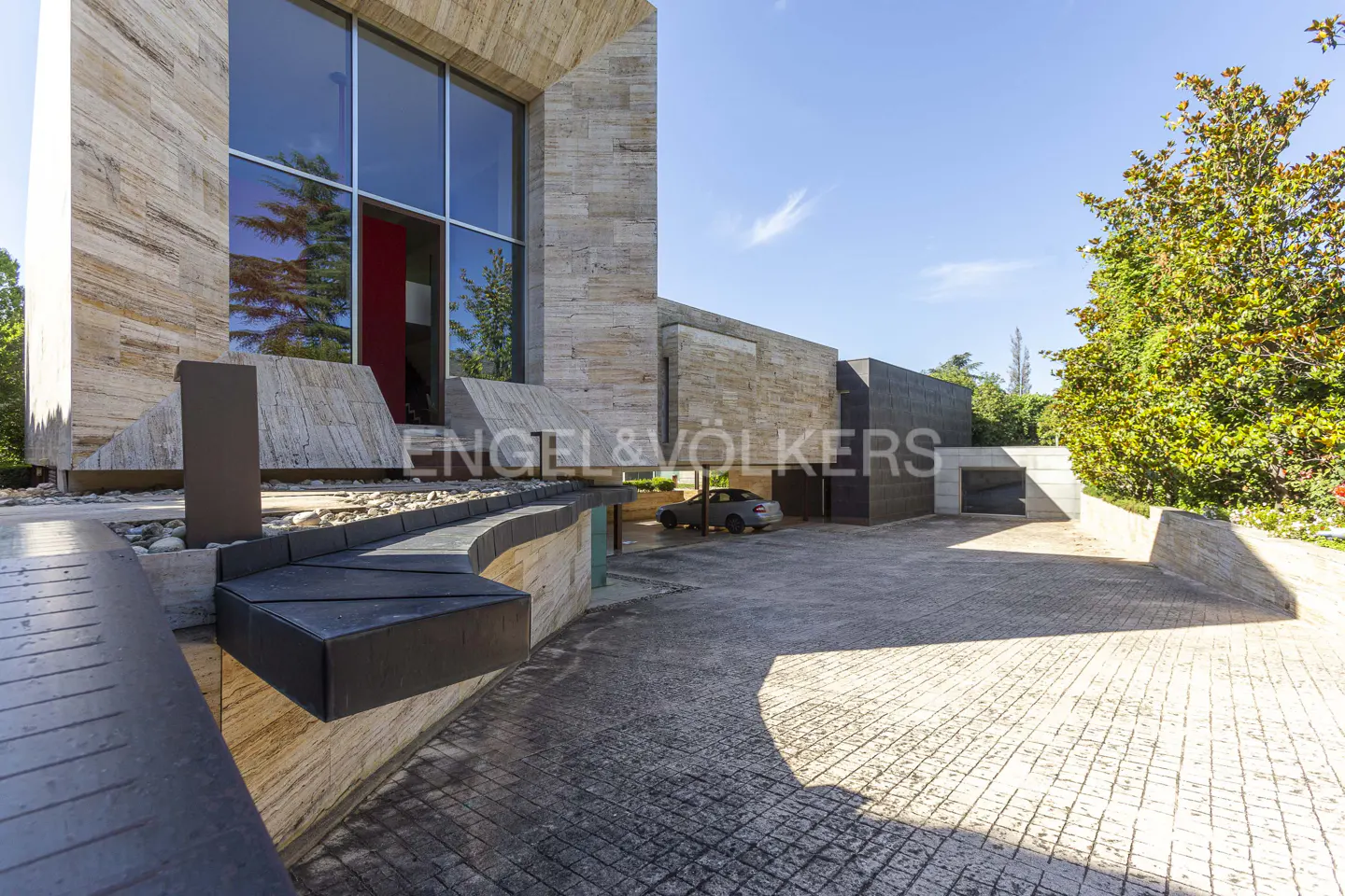 Exterior view of a modern stone house with a large window, driveway, and a car parked in the garage.
