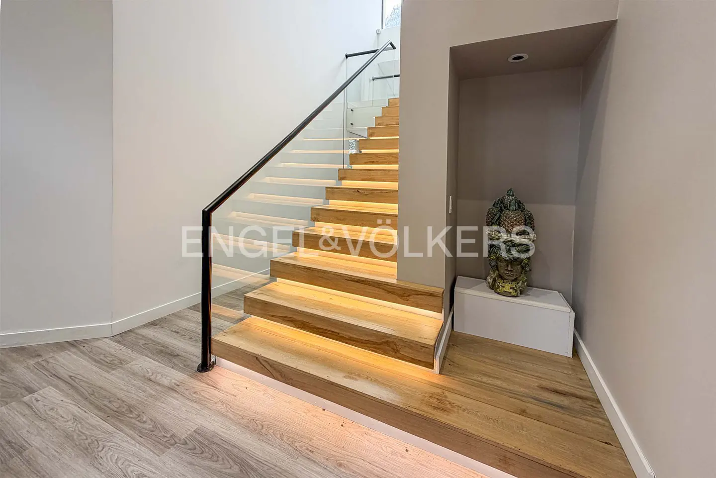 Modern staircase with light wood steps, glowing LED under-lighting, glass railing, and a decorative Buddha statue.