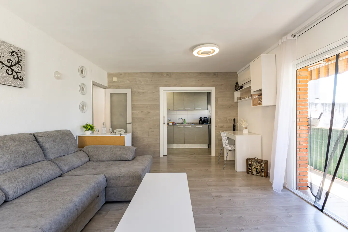 Modern living room with a grey sectional sofa, white coffee table, and a view into a kitchen with light wood-look walls.