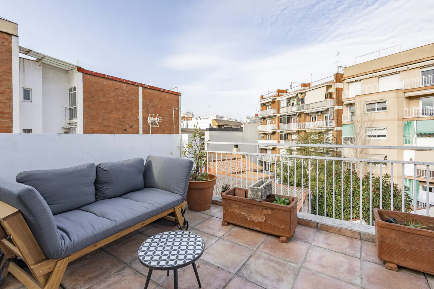 Rooftop patio with gray sofa, patterned table, and terracotta planters. White railing overlooks city buildings under a cloudy sky.
