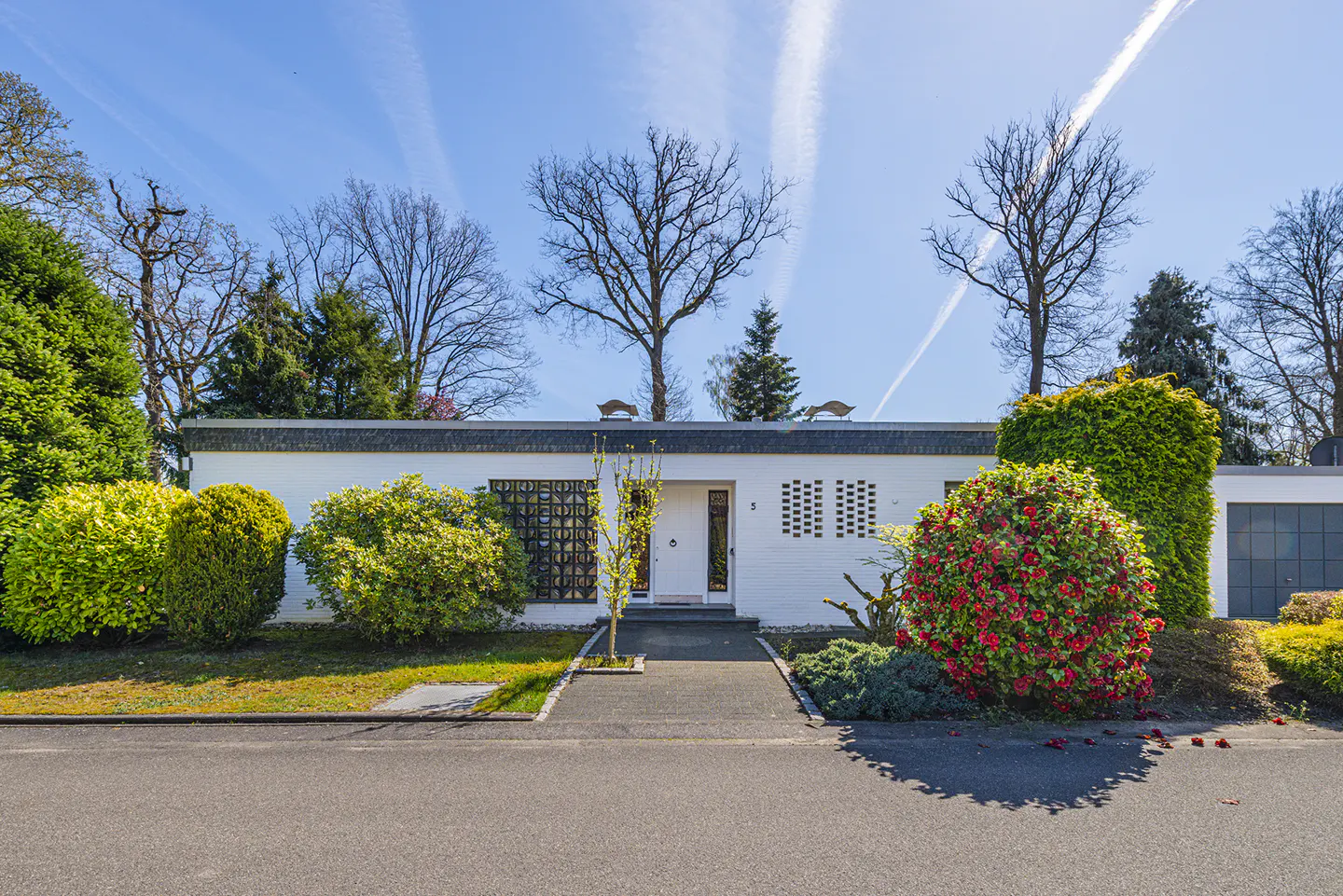 A modern, white, single-story house with a flat roof and manicured landscaping under a blue sky with contrails.
