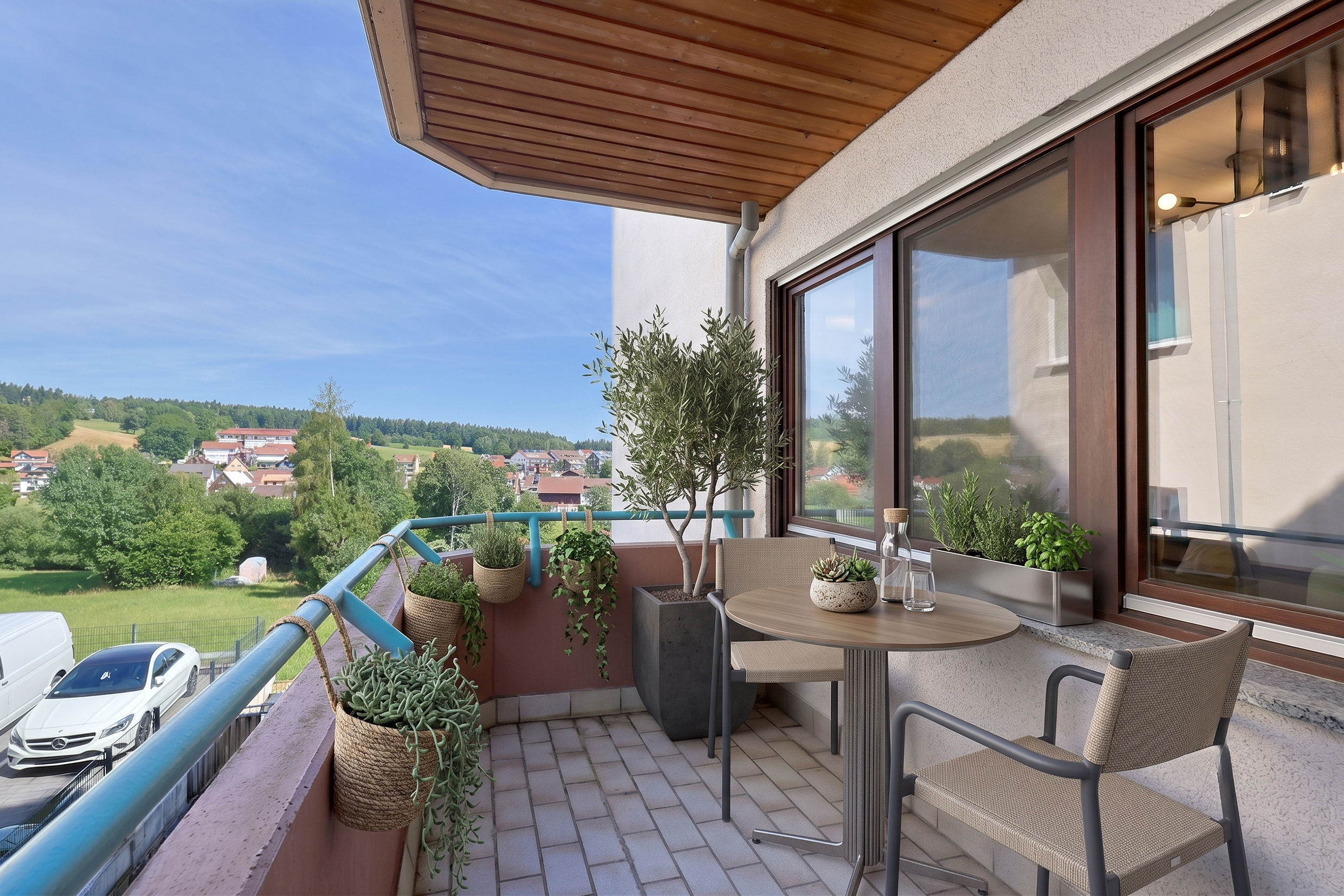 A balcony with a small round table, two chairs, potted plants, and a scenic view of a green landscape and distant houses.