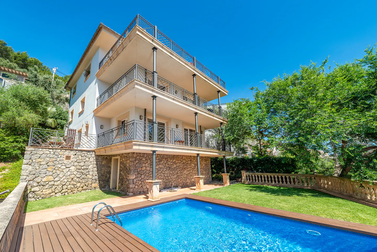 Three-story house with balconies overlooking a pool. The house has a stone base and is surrounded by green grass and trees.