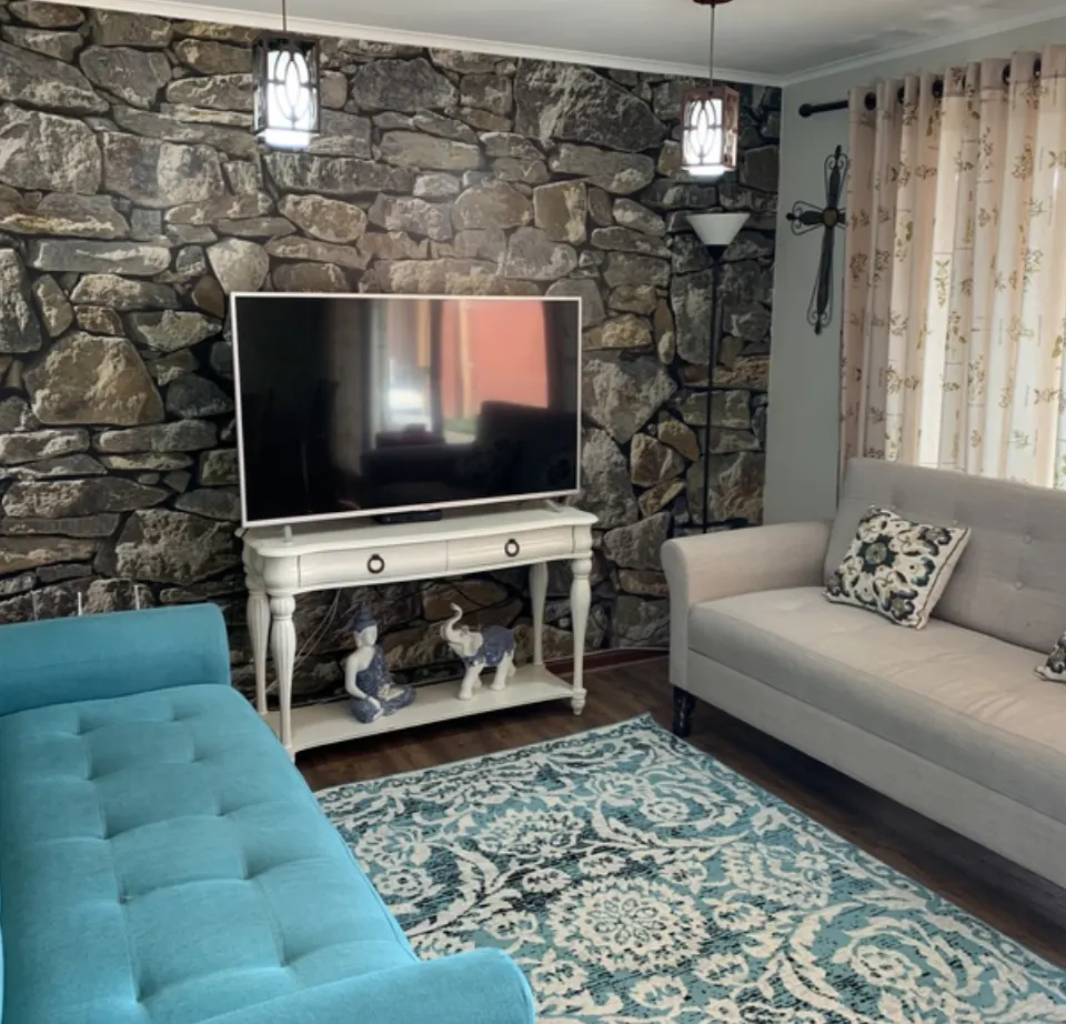 Living room with stone wall, TV on white console table, blue sofa, beige sofa, and patterned blue rug.