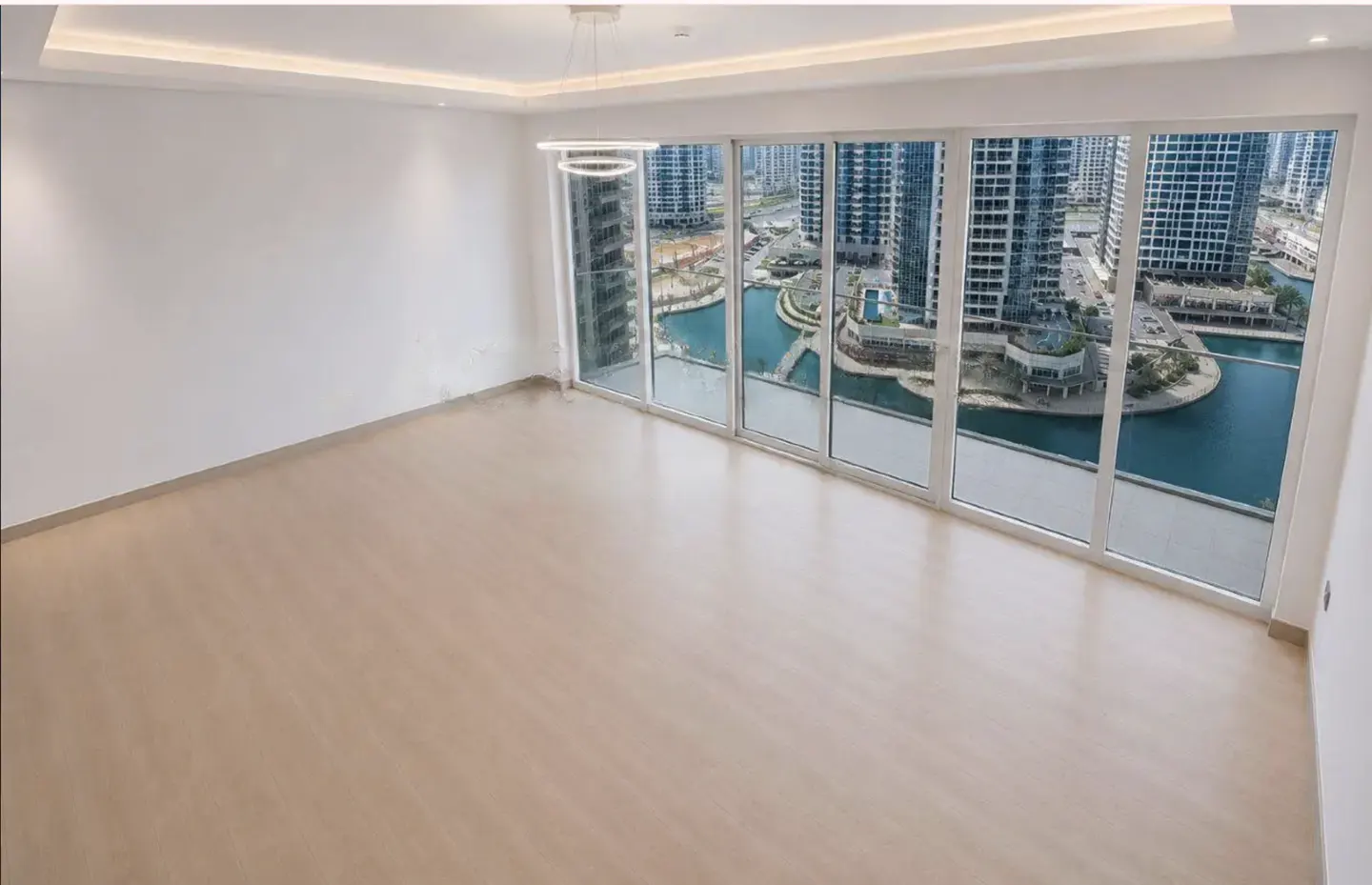 Empty room with light wood floors, white walls, and floor-to-ceiling windows overlooking a city canal and skyscrapers.