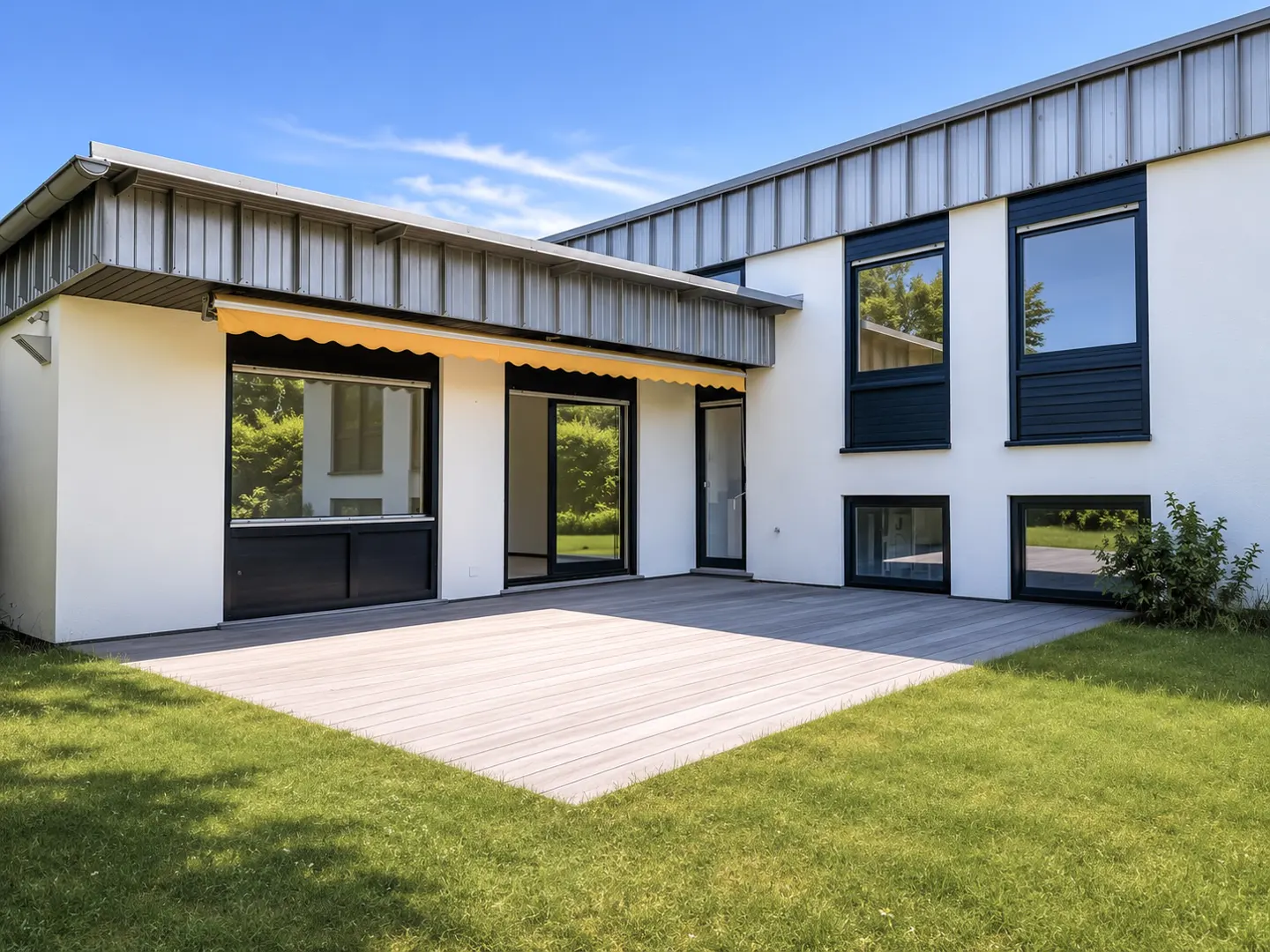 Modern white house exterior with a wooden deck, yellow retractable awning, and large black-framed windows facing a lawn.