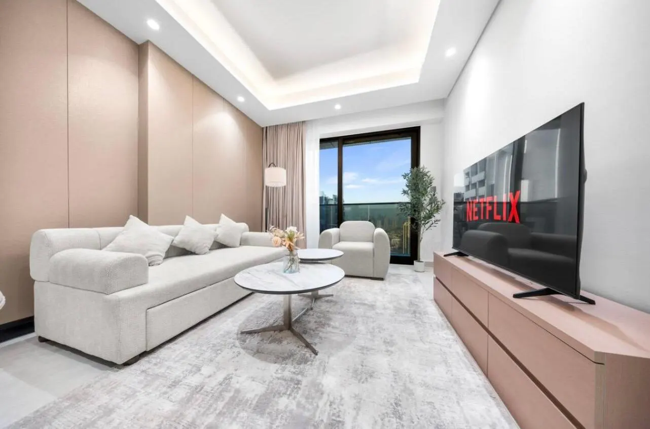 A modern living room with a white sofa, chair, and marble coffee table. A large TV displays the Netflix logo. Balcony view in the background.