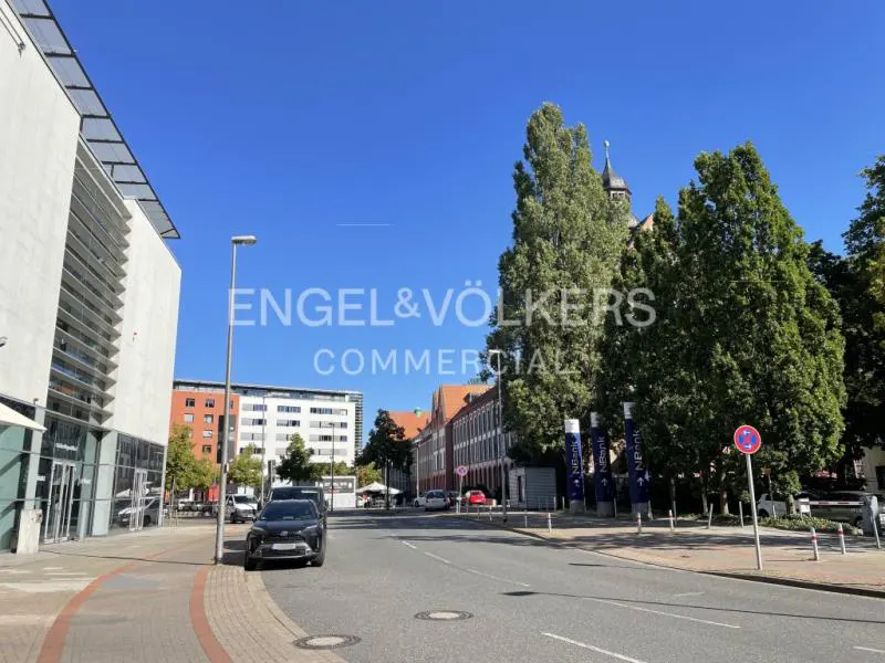 Street view with modern buildings, cars, and trees under a clear blue sky. An Engel & Völkers Commercial logo is superimposed on the image.