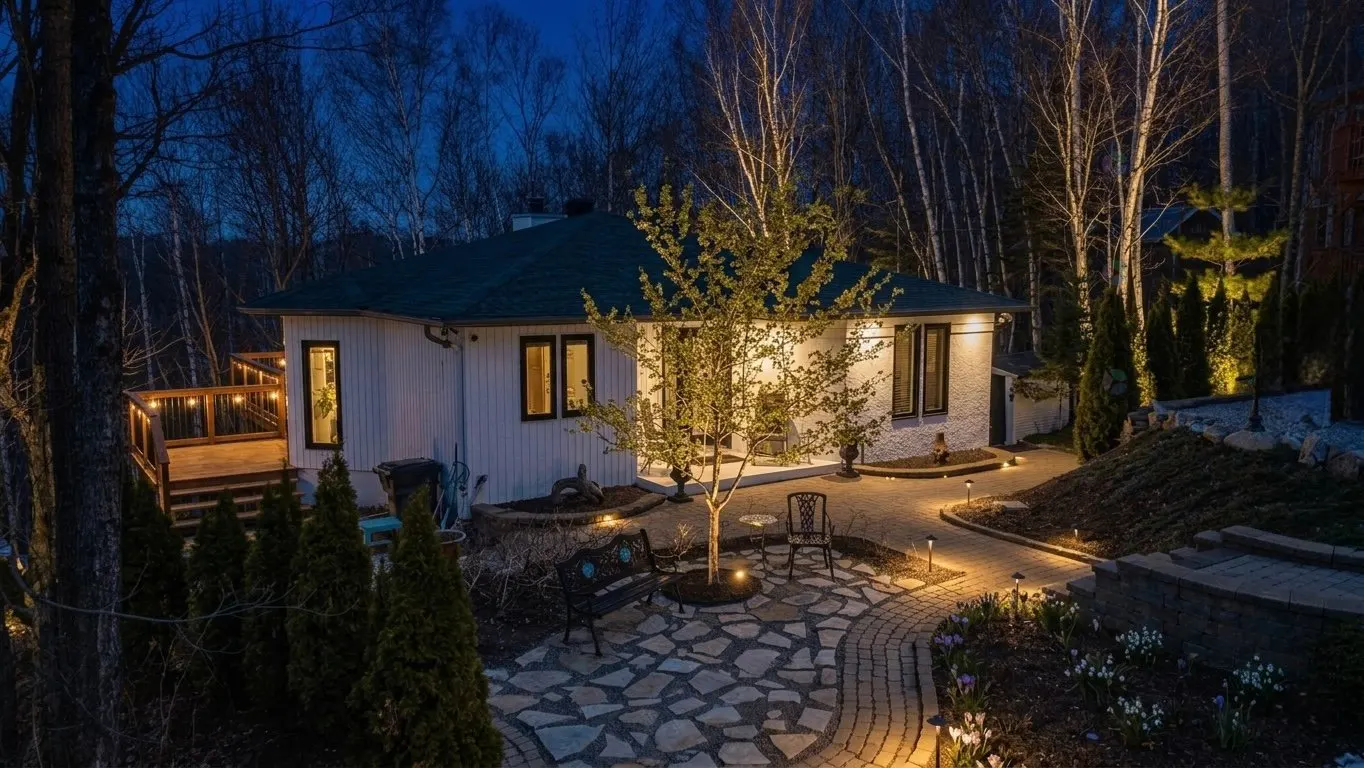 Night view of a white house with a green roof, surrounded by trees and illuminated landscaping. A stone patio features a bench and chairs.