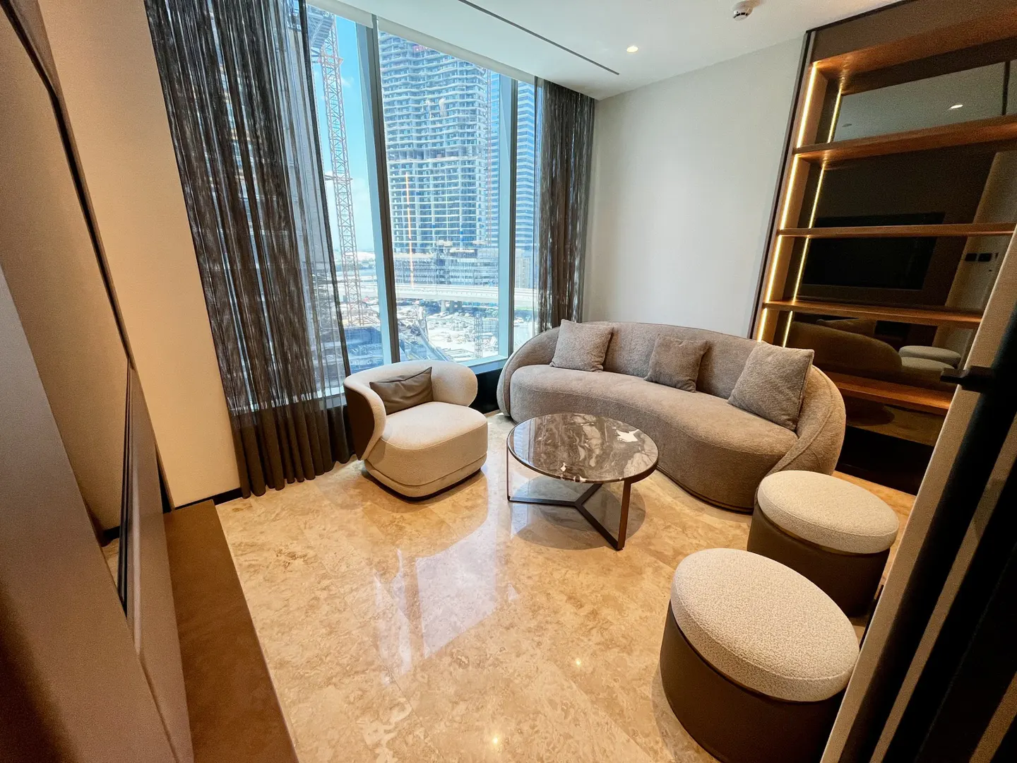 Living room with beige sofa, chair, and ottomans on a marble floor. A round coffee table sits in the center, with a city view visible through the window.