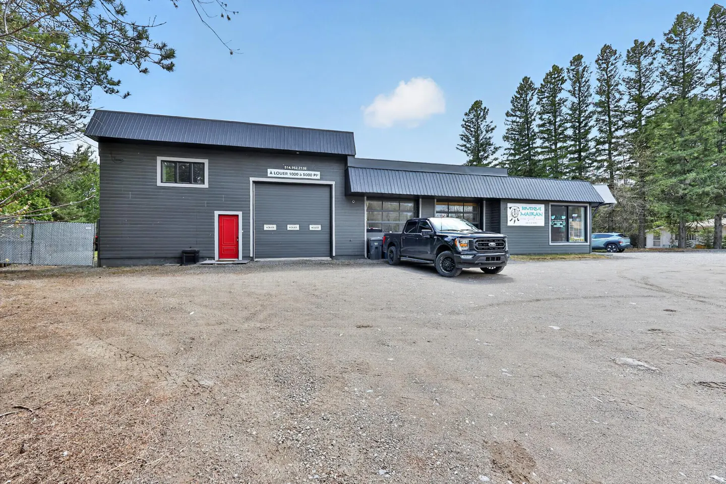 Gray commercial building with a red door, garage door, and a black pickup truck parked in front. Trees in the background.