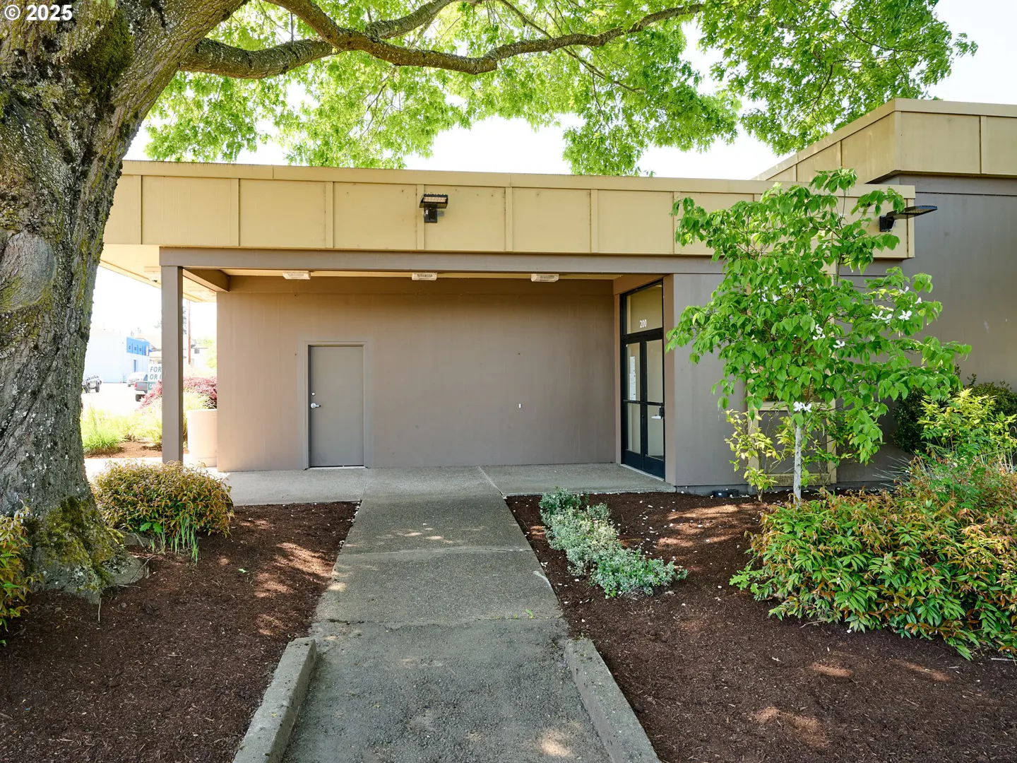 Exterior of a tan building with a covered entrance, a gray door, and a black-framed glass door. A tree is on the left and landscaping is in front.