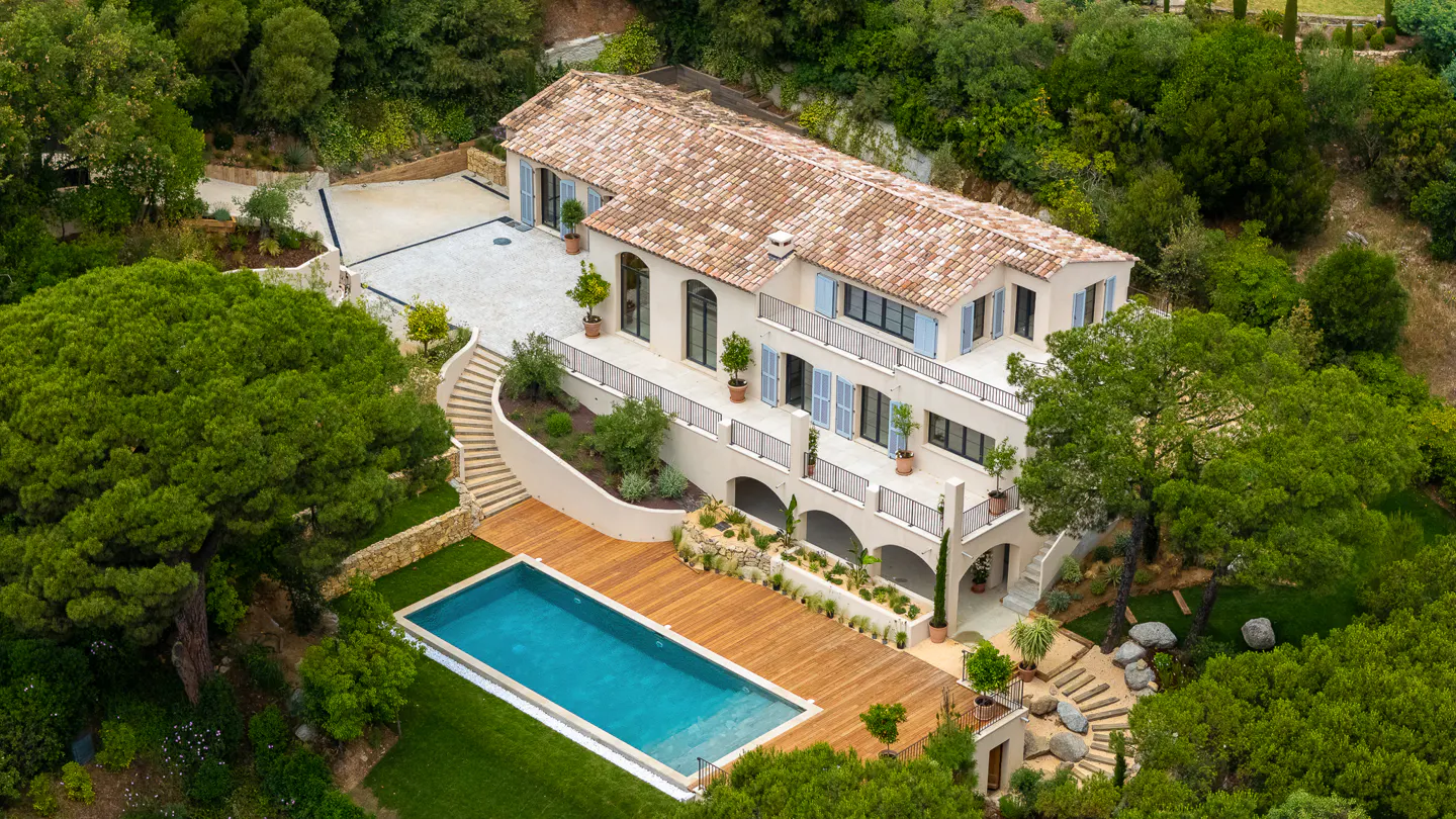 Aerial view of a multi-story beige house with a terracotta tile roof, blue shutters, a pool, and lush green trees.