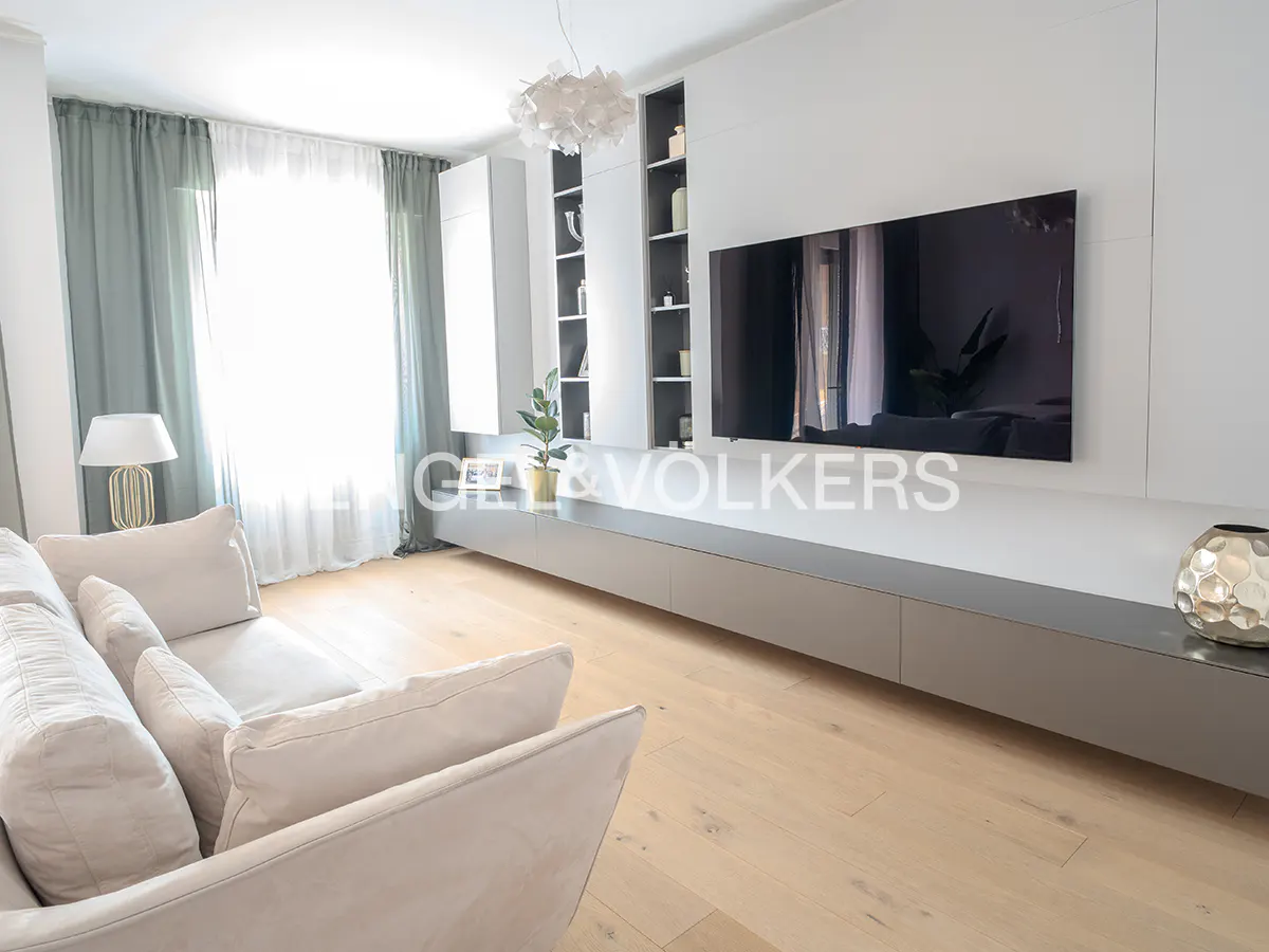 Bright living room with a beige sofa, light wood floors, and a large TV mounted on a white wall unit. Green curtains frame a window.