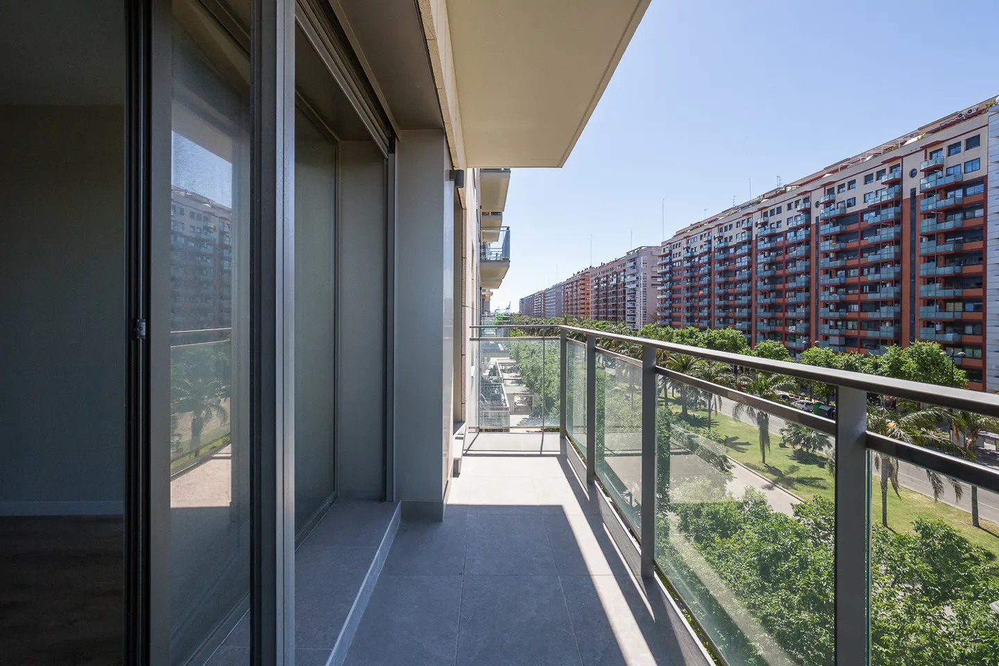 Apartment balcony with glass railing overlooking a tree-lined street and tall buildings under a clear blue sky.