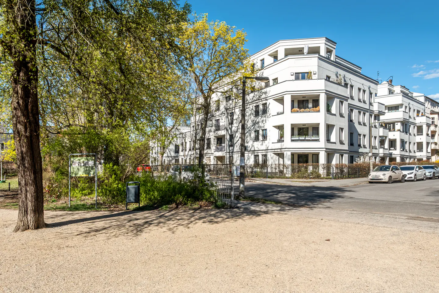 Modern white apartment building with balconies, seen from a park with trees and gravel path on a sunny day.