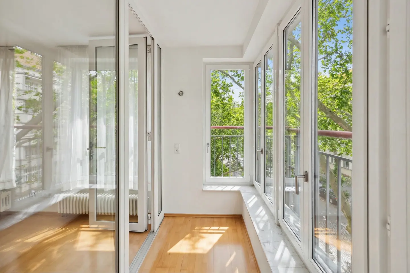 Bright sunroom with wood floors and white walls. Large windows offer a view of green trees and a blue sky.