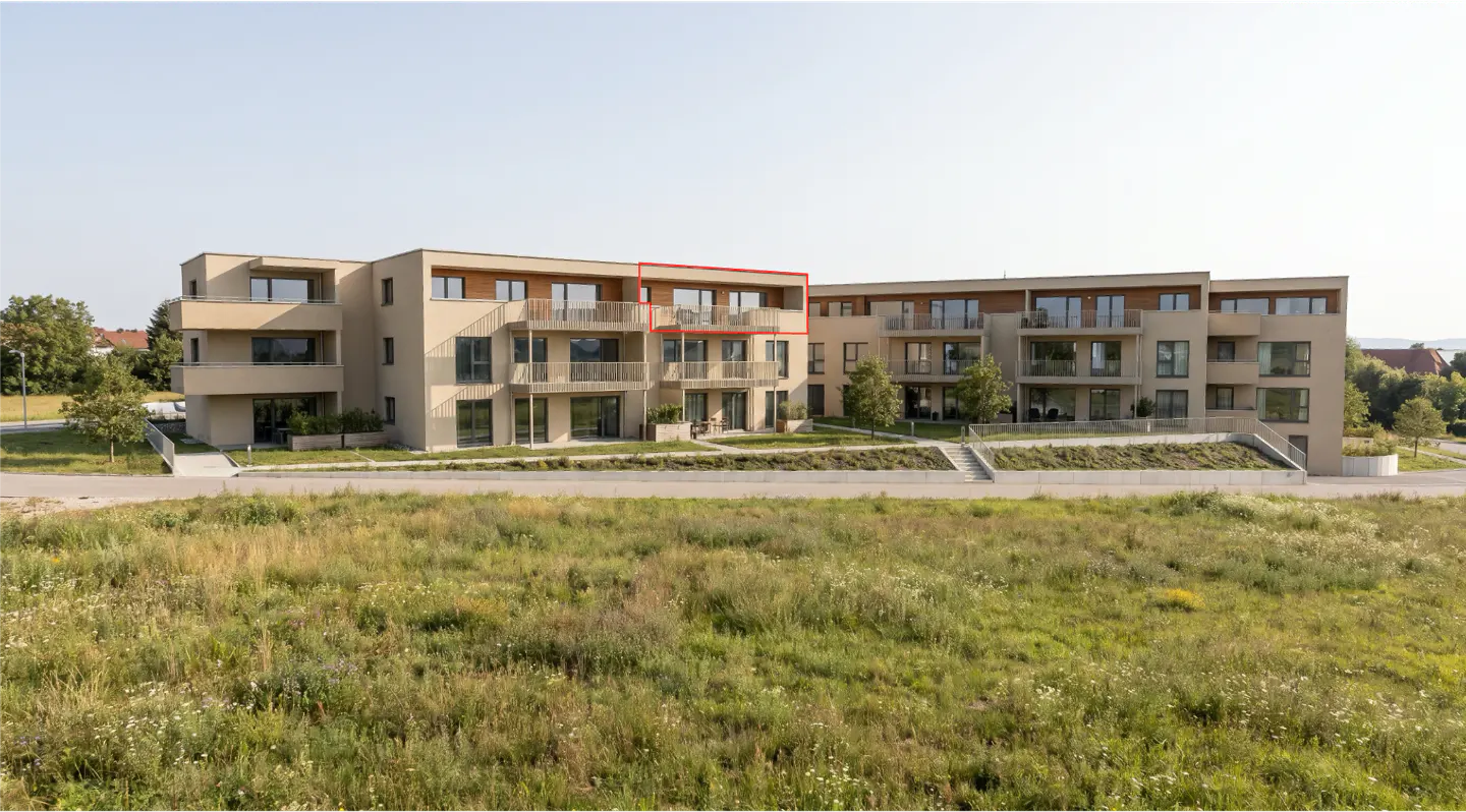 Modern beige apartment complex with balconies, set against a grassy field, with one top-floor unit outlined in red.