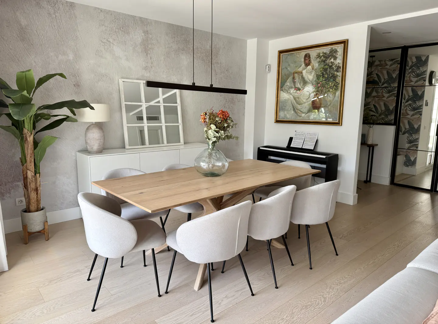 A bright dining room with a light wood table, white chairs, and a black piano in the background.