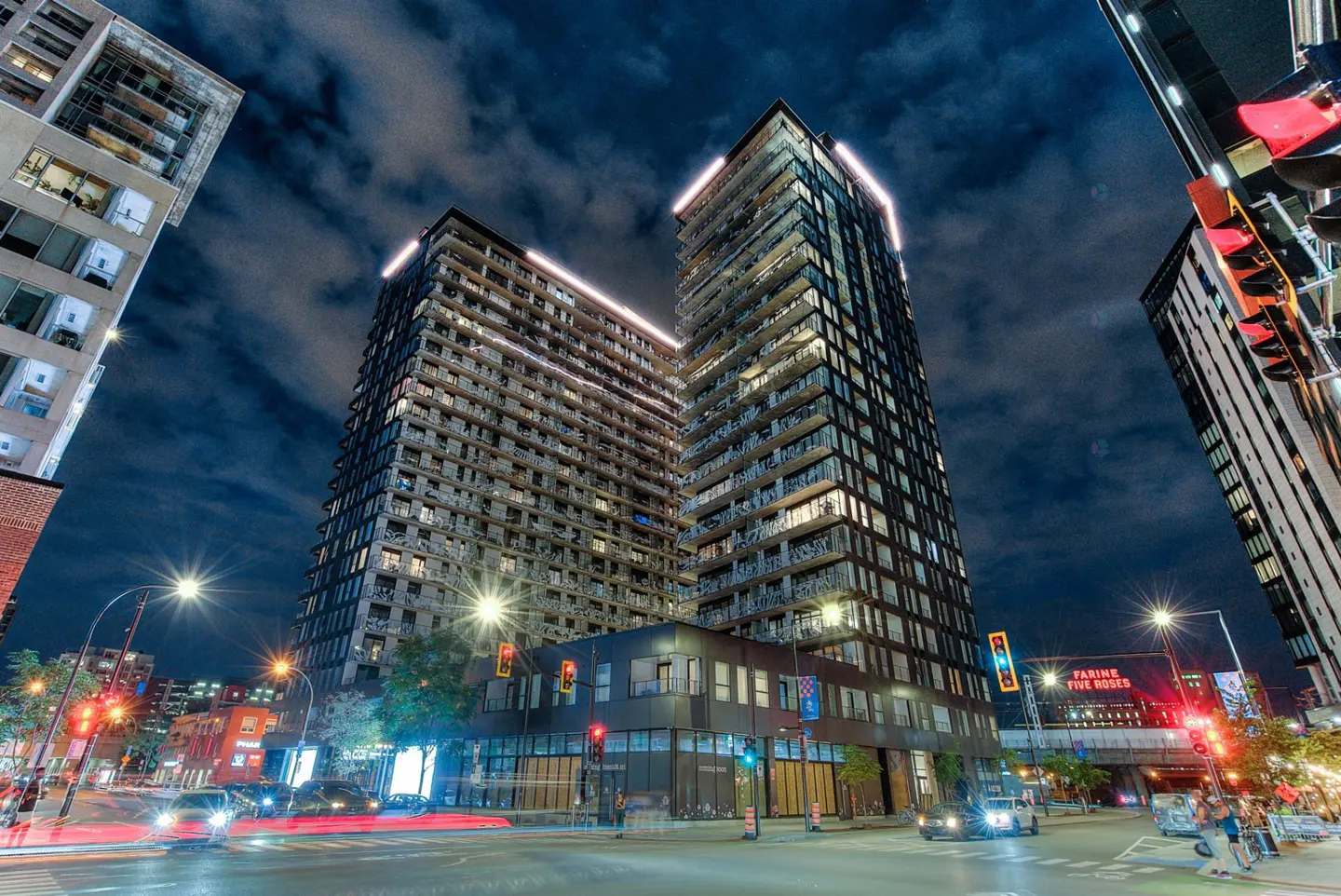 Night view of two modern high-rise apartment buildings with illuminated tops, street with cars, and city lights.