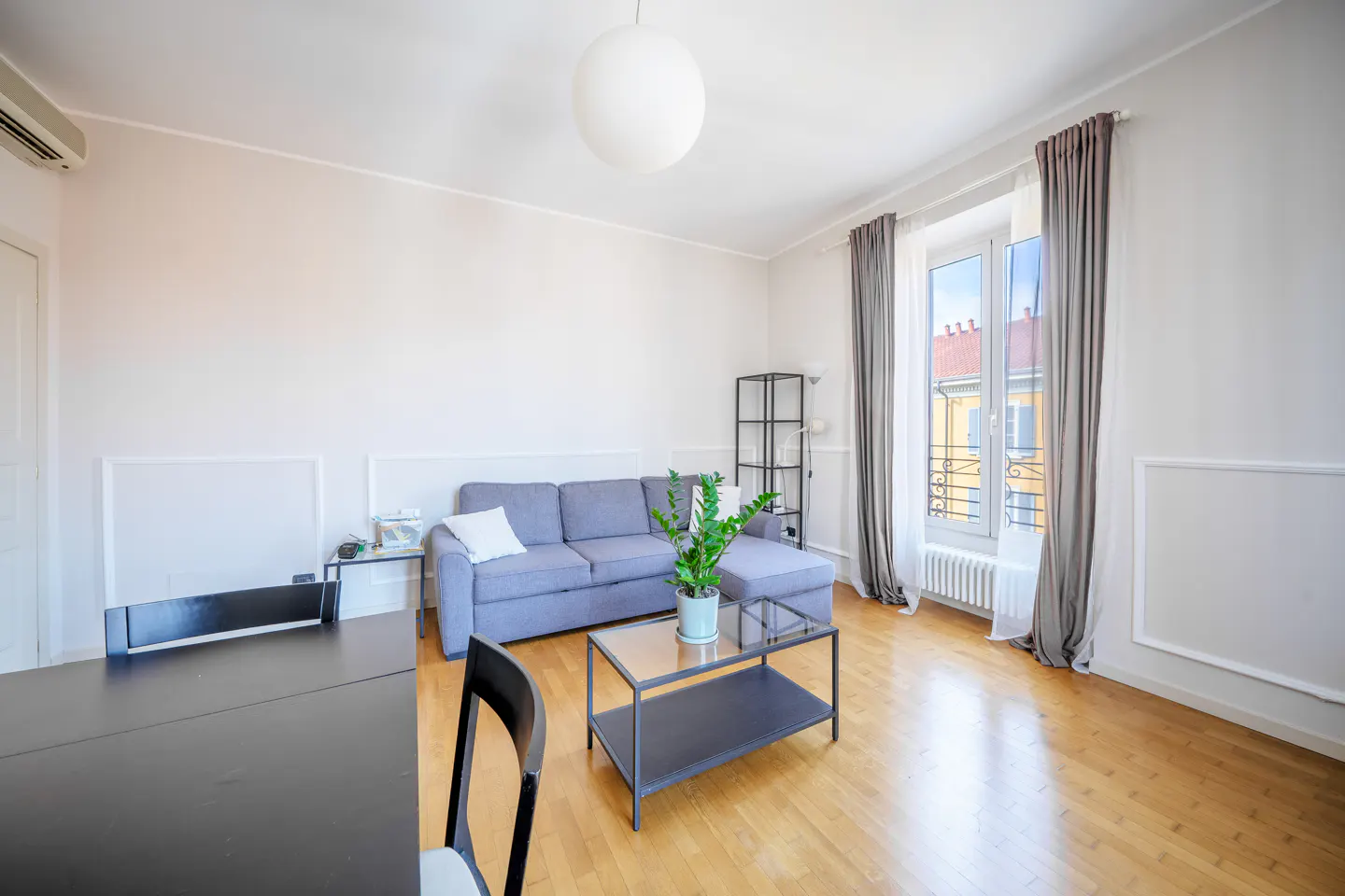 Bright living room with a gray sofa, black table, wood floors, and large window with gray curtains.