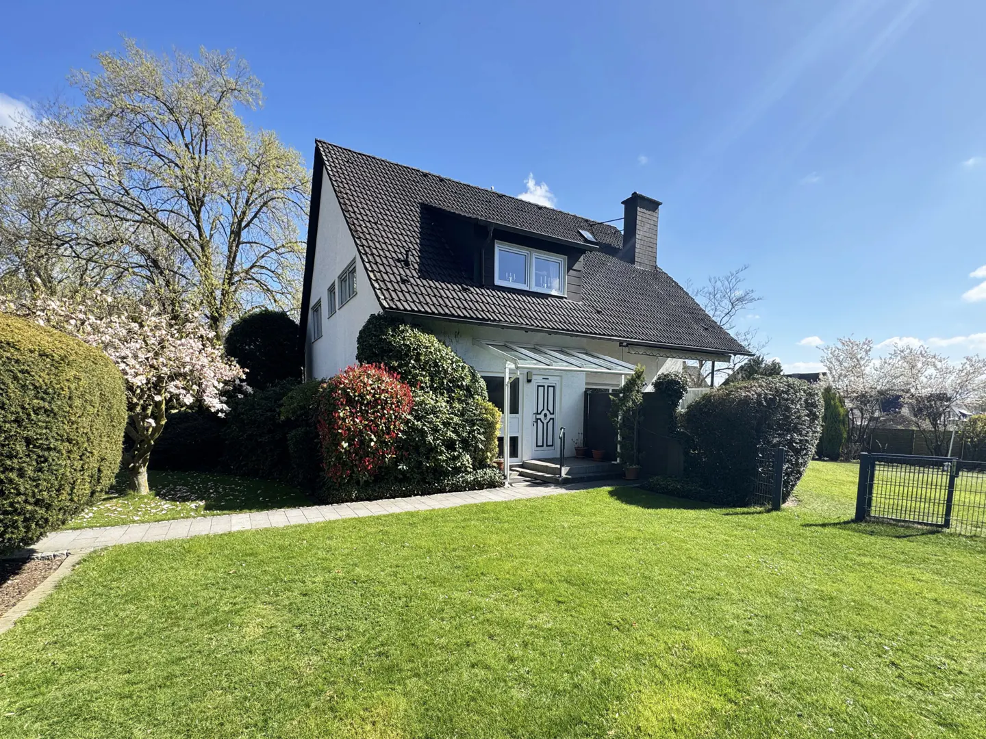 A white house with a dark roof sits on a green lawn under a blue sky. Bushes and trees surround the house.