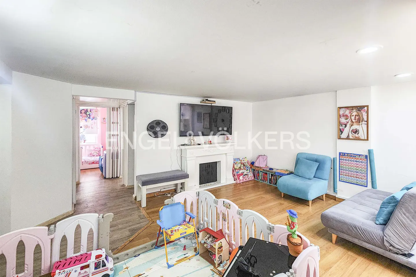 Bright, open-plan living room with a play area. Features a TV over a white fireplace, blue sofa and chair, and a child's playpen with toys.