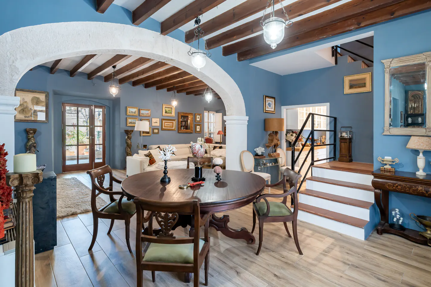 Dining area with a round wooden table, green-cushioned chairs, blue walls, exposed ceiling beams, and a stone archway.